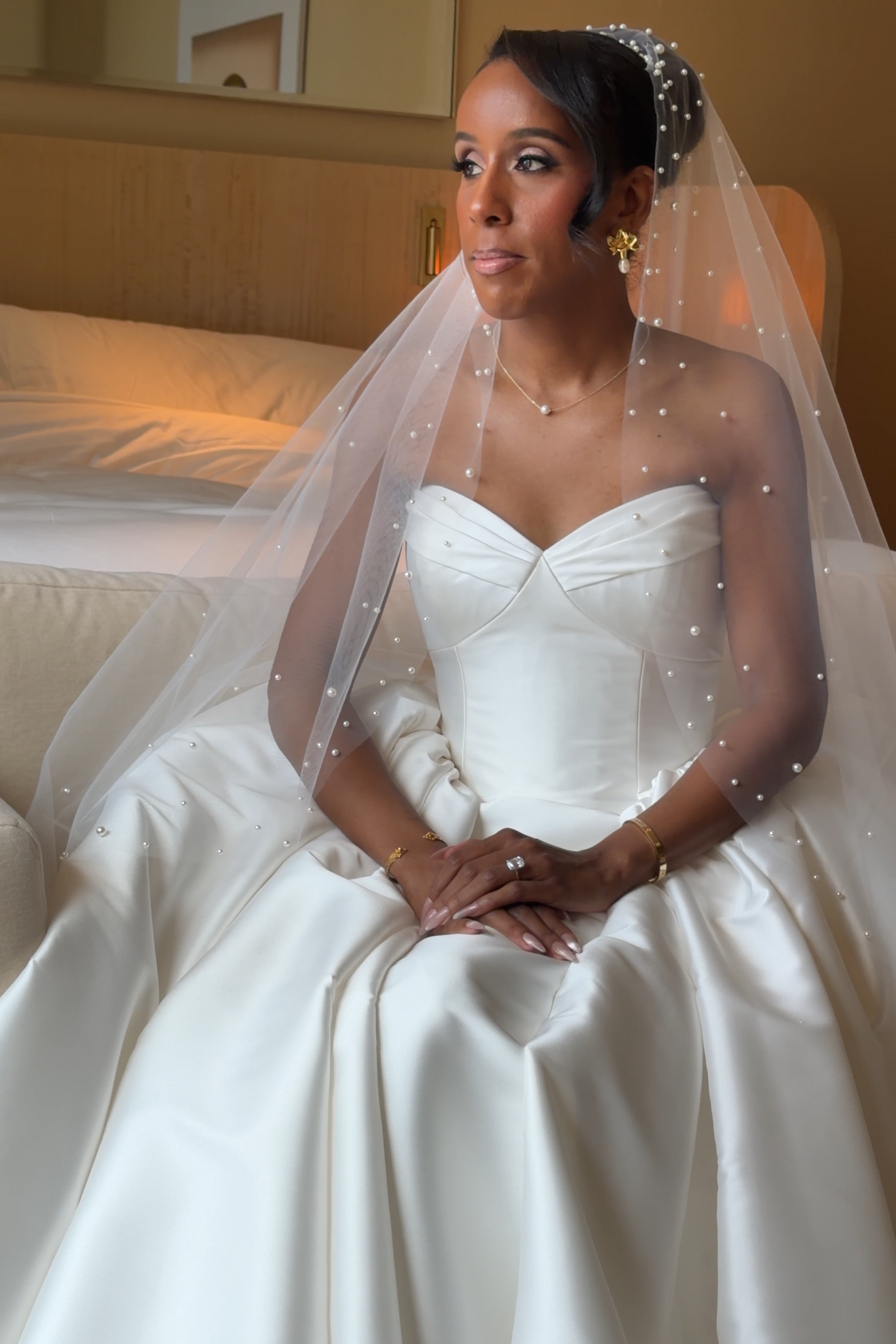 Bride sitting on a bed in a hotel room, wearing a white wedding dress, veil with pearl embellishments, and gold jewelry, looking slightly to her left.