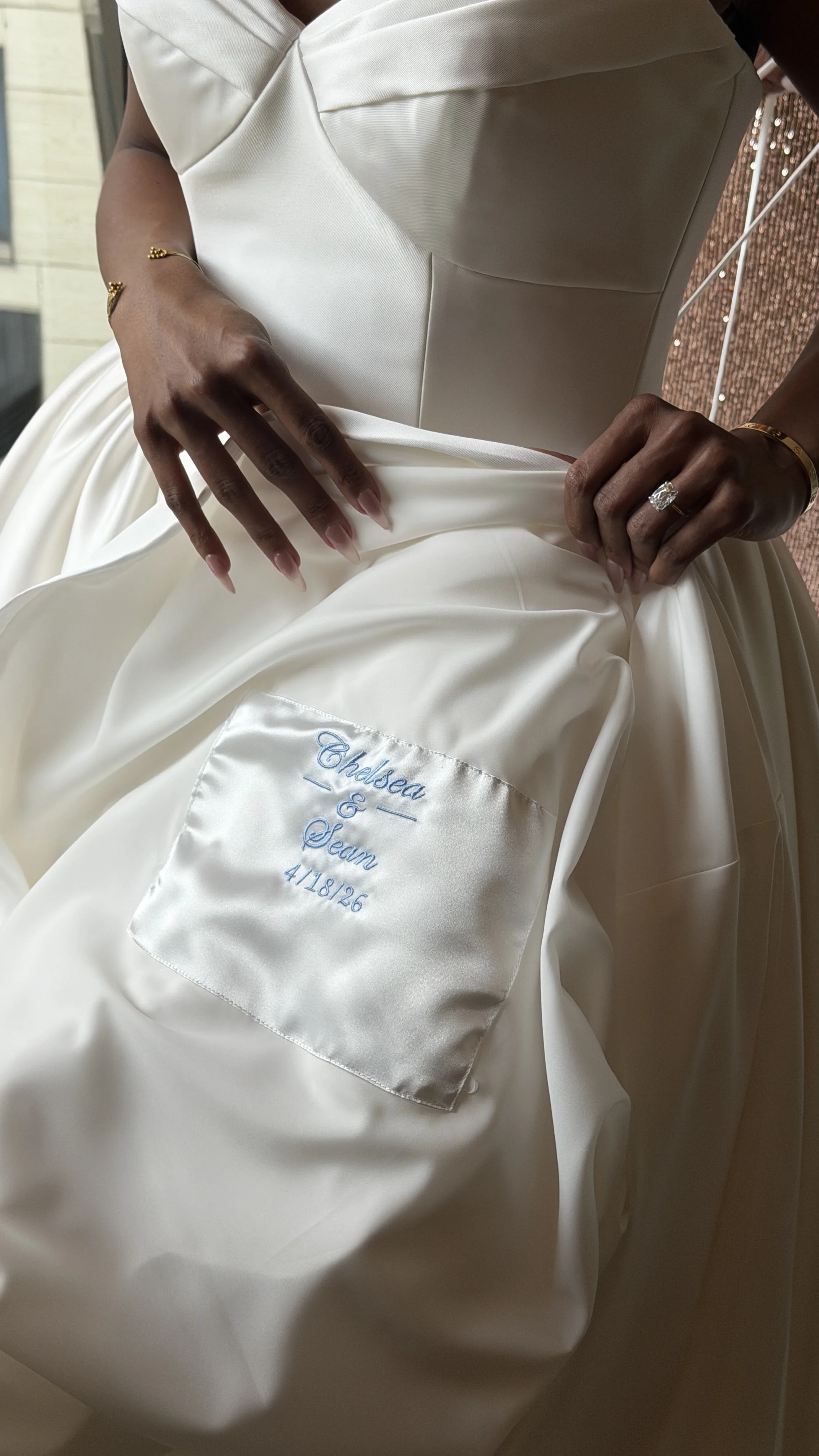 Close-up of a person in a white wedding gown, holding folded fabric with a satin pocket embroidered with blue text reading 'Chalsea & Seann 4/18/26'.