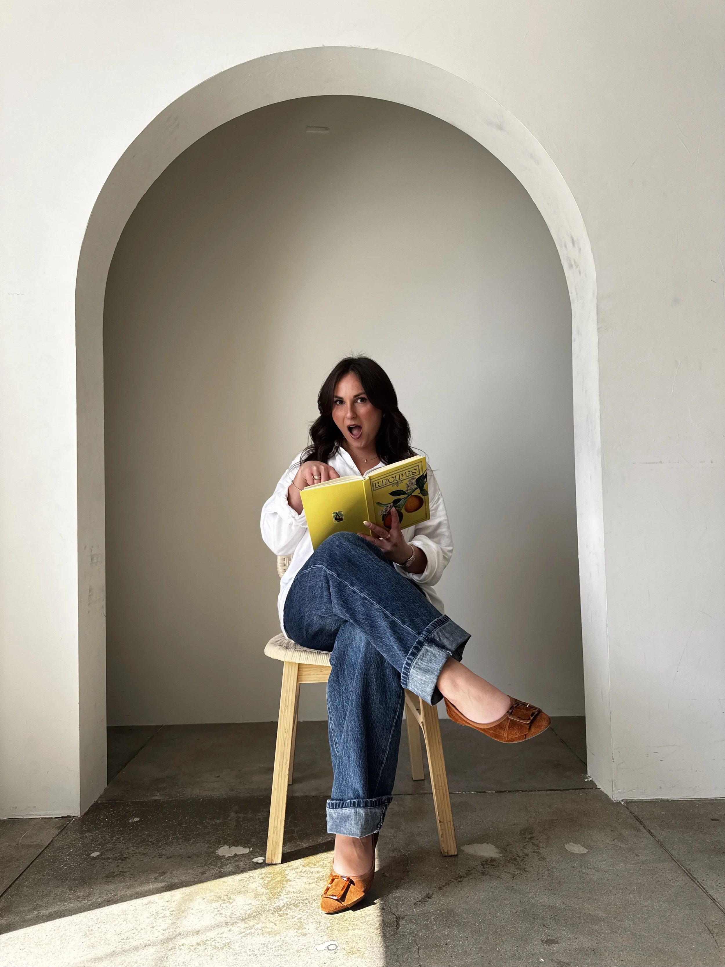 A woman sitting on a stool reads a yellow cookbook titled "Recipes" with an expression of surprise or excitement, in a minimalist white room with an arched niche.