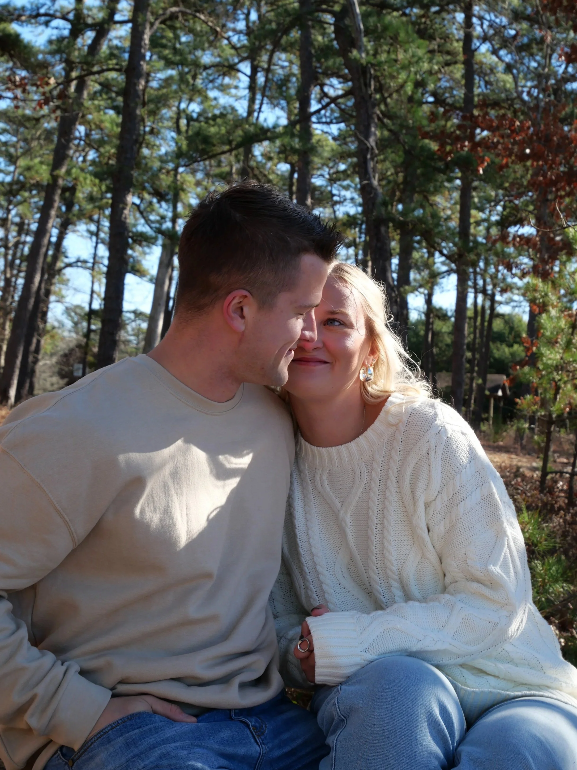 A young man and woman sit close together outdoors in a wooded area, smiling and looking at each other affectionately.