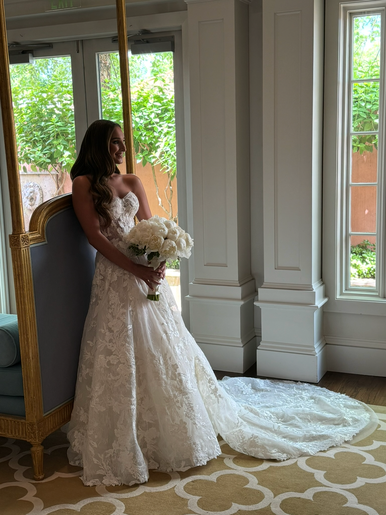 A smiling bride in a white lace wedding dress holding a bouquet of white flowers, standing indoors near a large mirror and window with greenery outside.