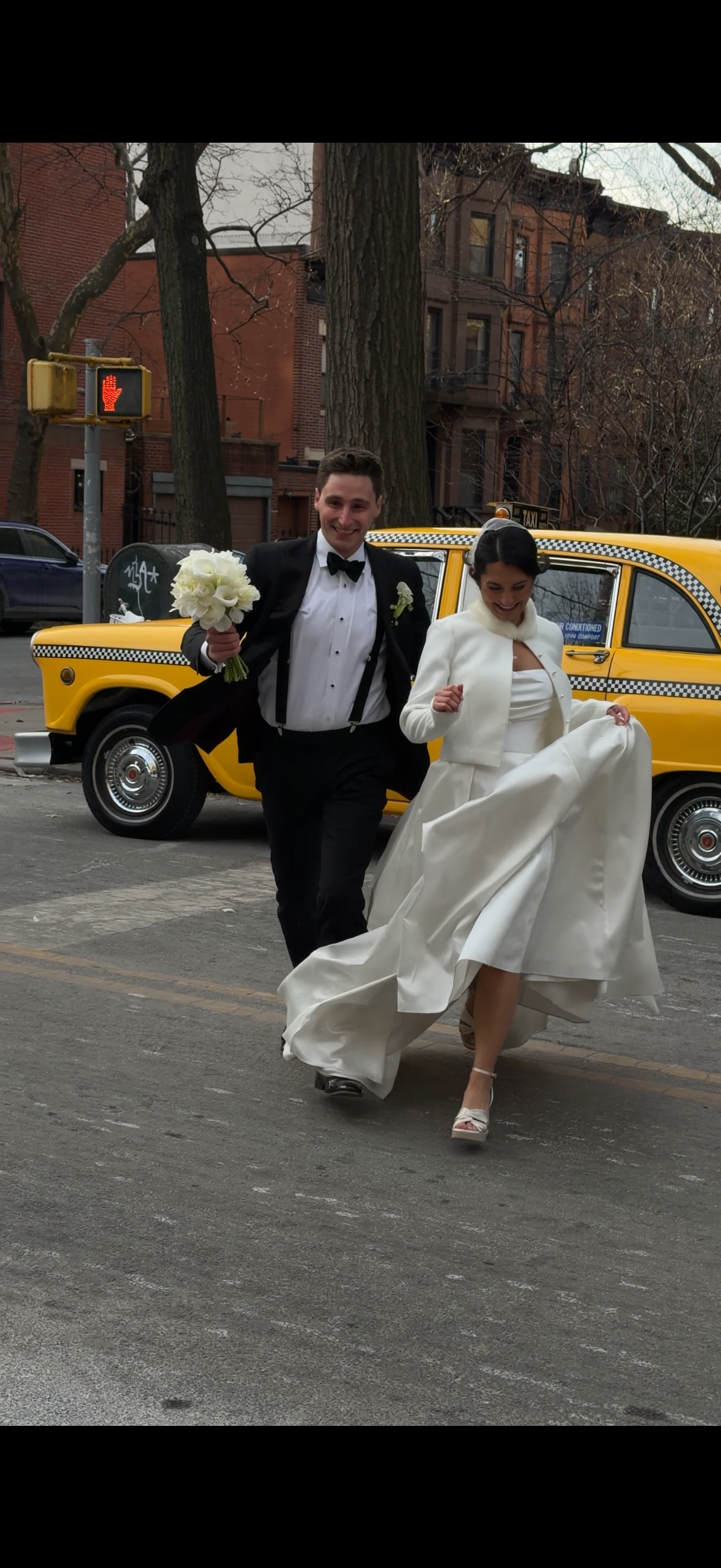 A bride and groom running on a city street, with the bride holding her wedding dress and the groom holding a bouquet of white flowers, in front of a yellow taxi cab.
