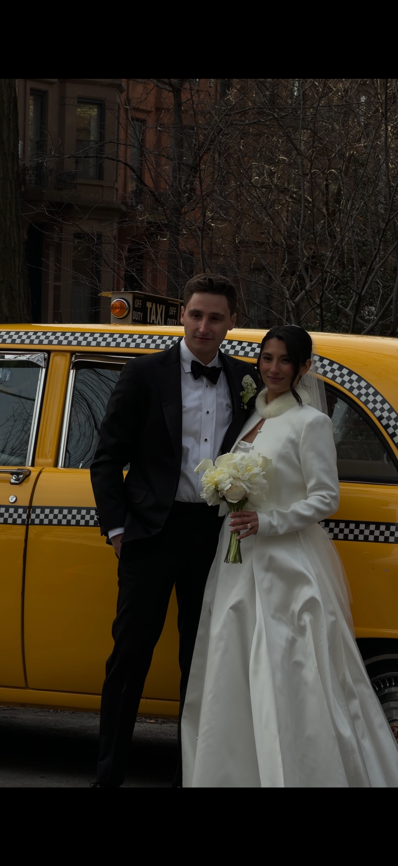 A newlywed couple standing in front of a yellow taxi cab, with the bride in a white wedding gown holding a bouquet of white flowers, and the groom in a black tuxedo.