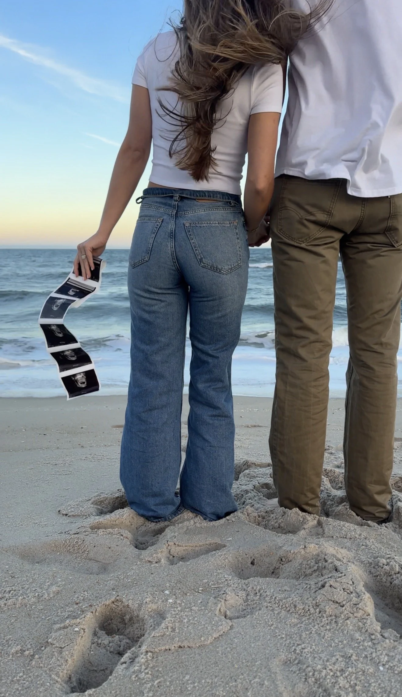 Two people are standing on the sandy beach holding hands, with footprints in the sand in front of them, and a calm ocean in the background during sunset or sunrise.