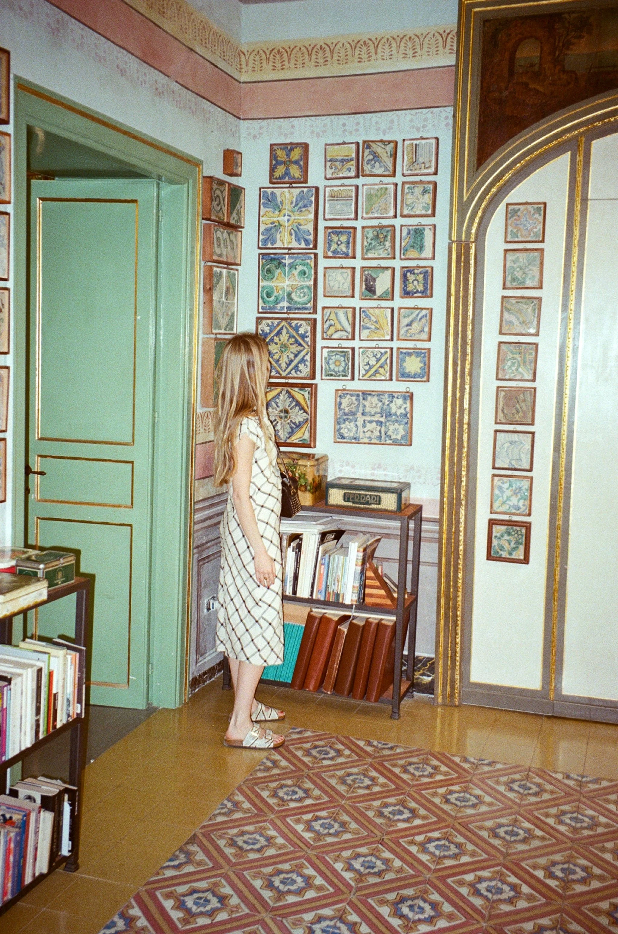 A woman with long blonde hair wearing a beige dress with black checkered pattern and white sandals is standing in a room filled with colorful, decorative tiles on the wall and a patterned carpet on the floor. There are bookshelves, a small table, and framed images on the walls.