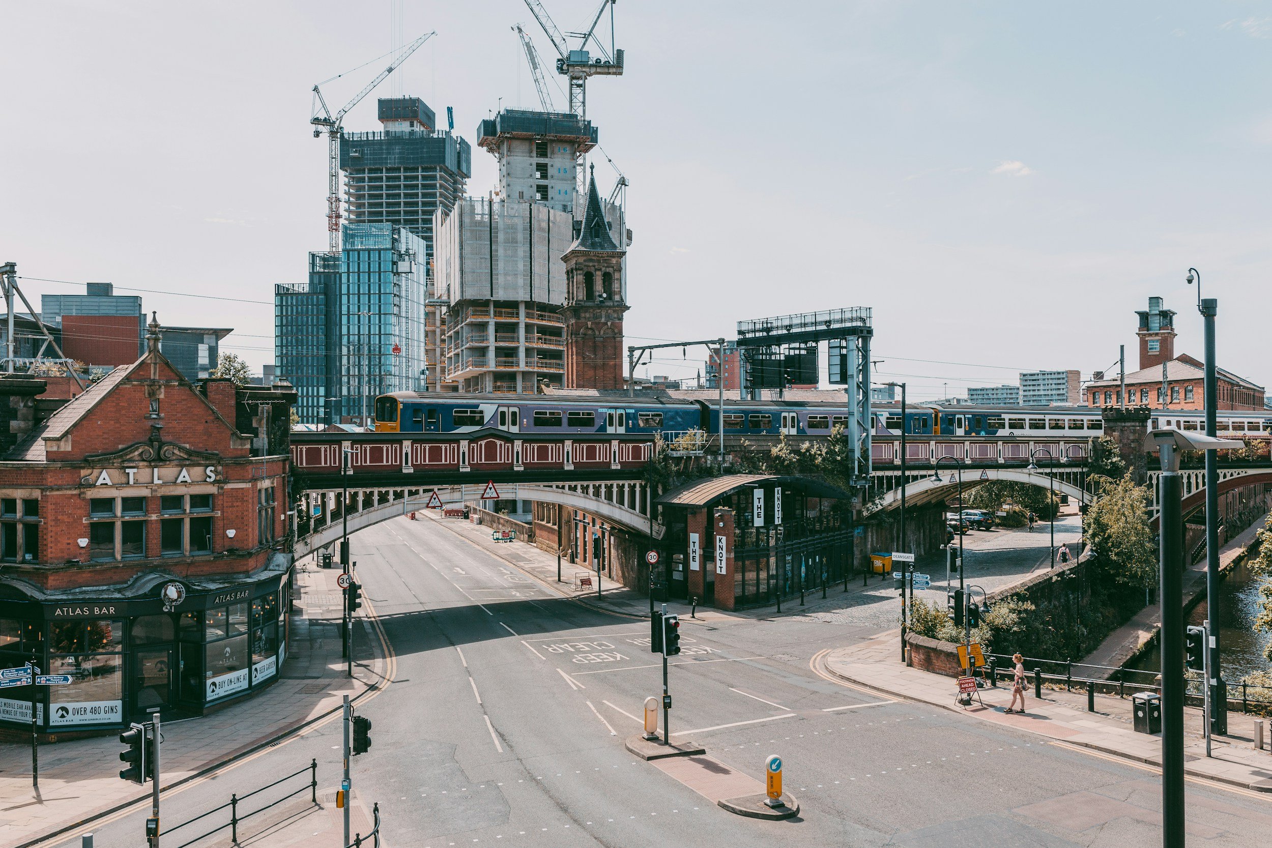 A cityscape featuring a mix of old brick buildings, a church with a clock tower, and modern skyscrapers under construction in the background. There is a railway with a train on an overpass, and empty streets with traffic lights and a few pedestrians.