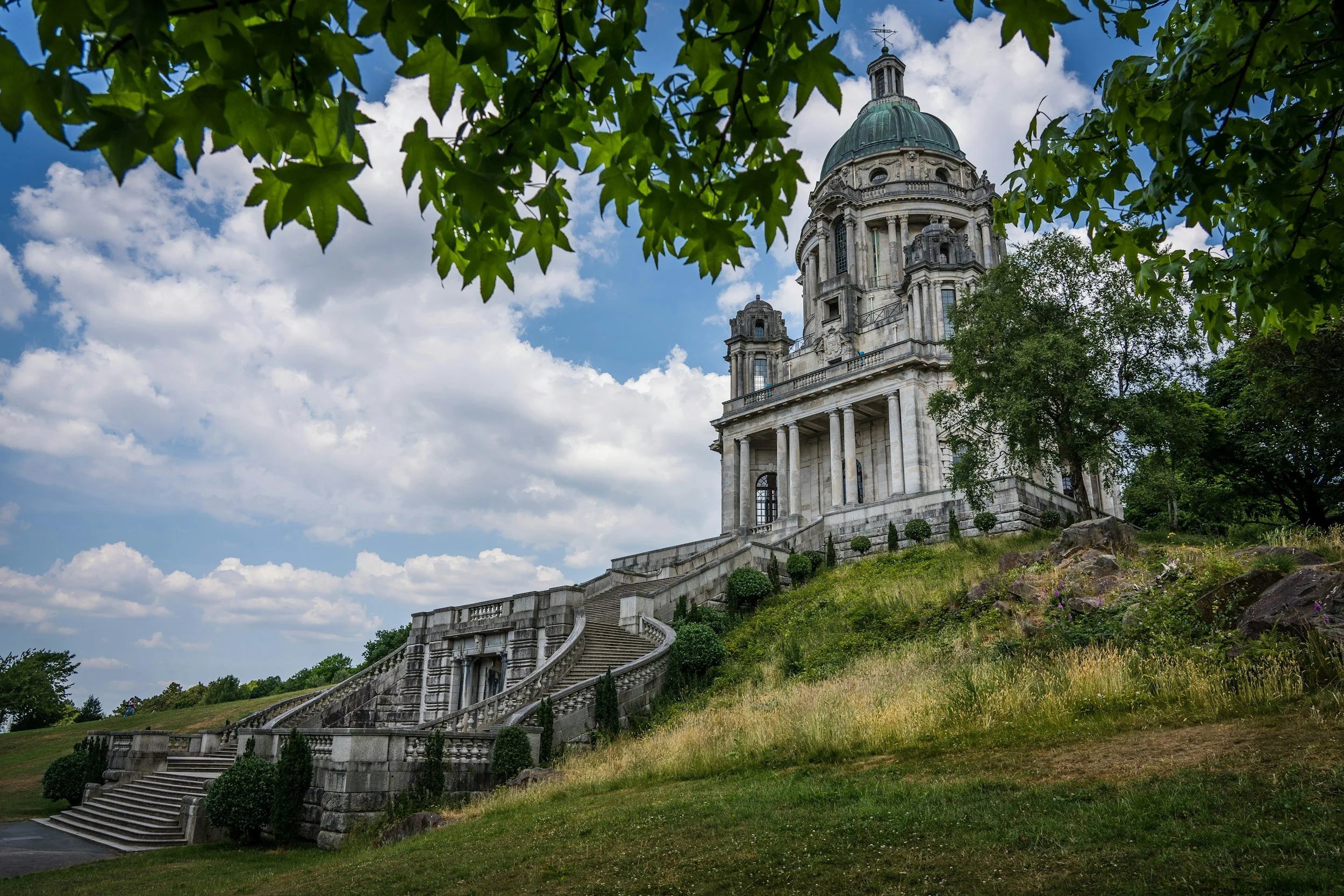A historic stone building with a domed roof, overlooking a grassy area with steps and surrounded by trees, under a partly cloudy sky.