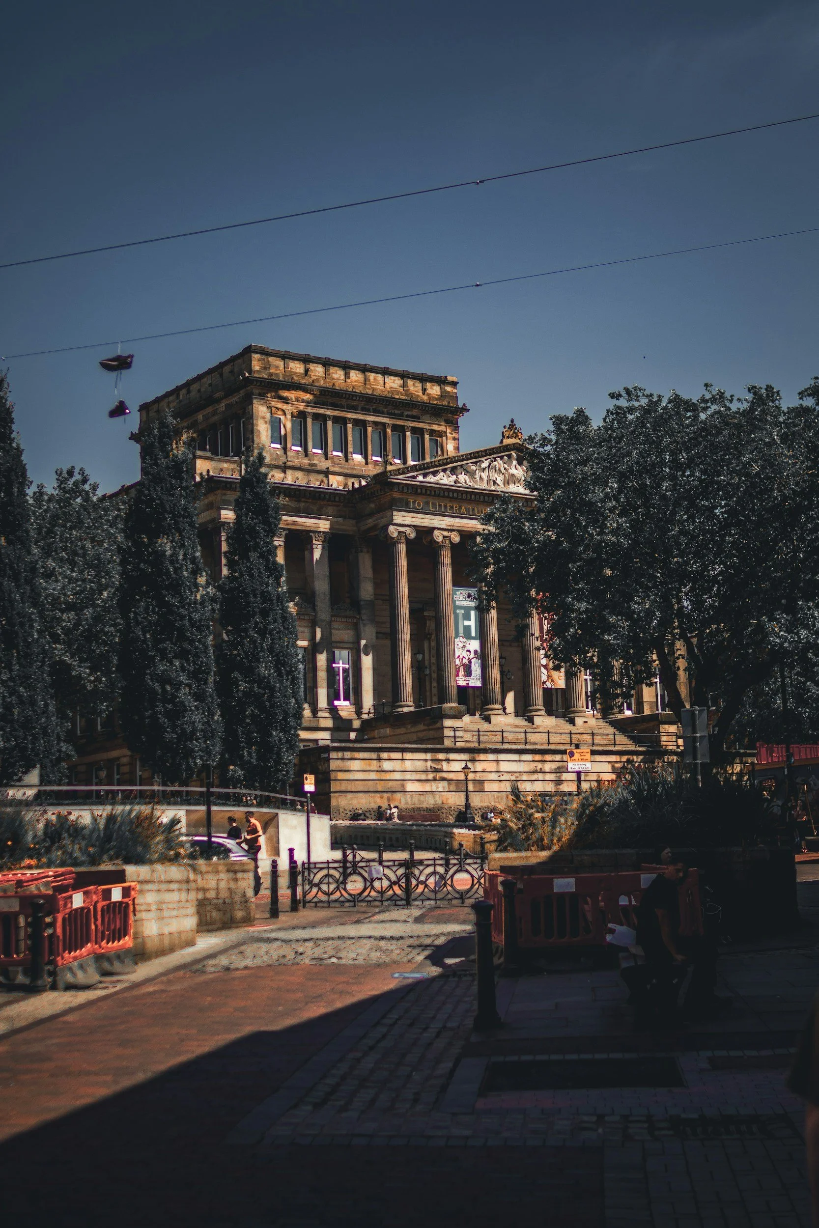 A historic building with classical architecture including columns, atop a flight of stairs, with trees and street scenery in front, under a clear sky with power lines overhead.