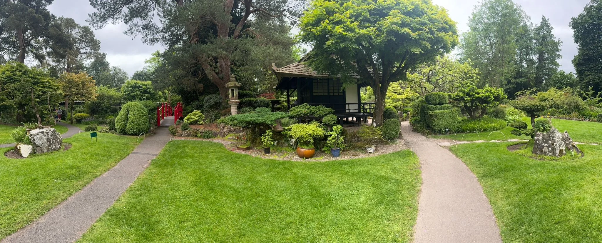 A tranquil Japanese-style garden with manicured trees and shrubs, a small pavilion, a red arched gateway, stone lanterns, and multiple walking paths.