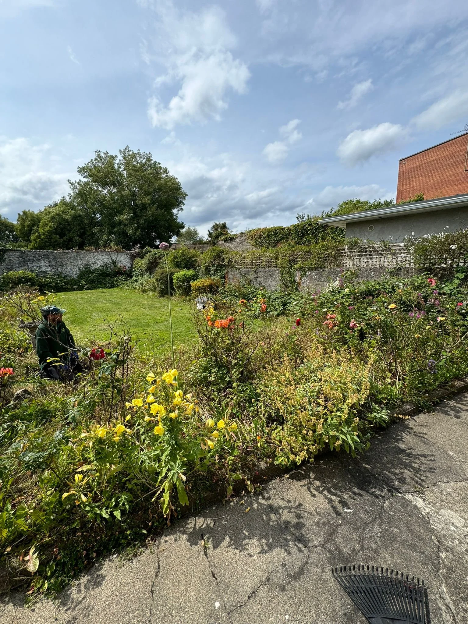 A backyard with a grassy lawn, flower garden, stone wall, and a partly cloudy sky.