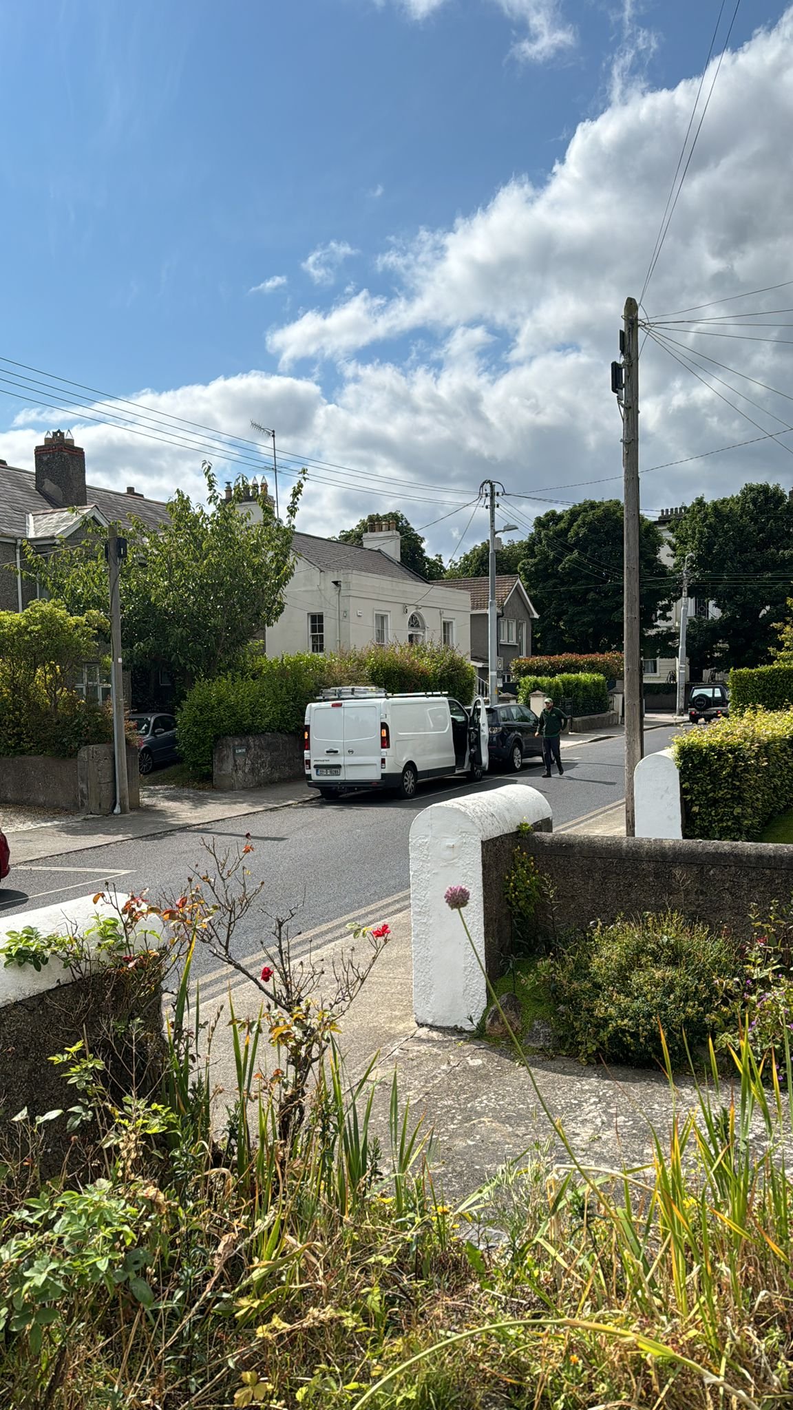 Residential street scene with white houses, parked cars, a person standing on the sidewalk, trees, and a blue sky with clouds.