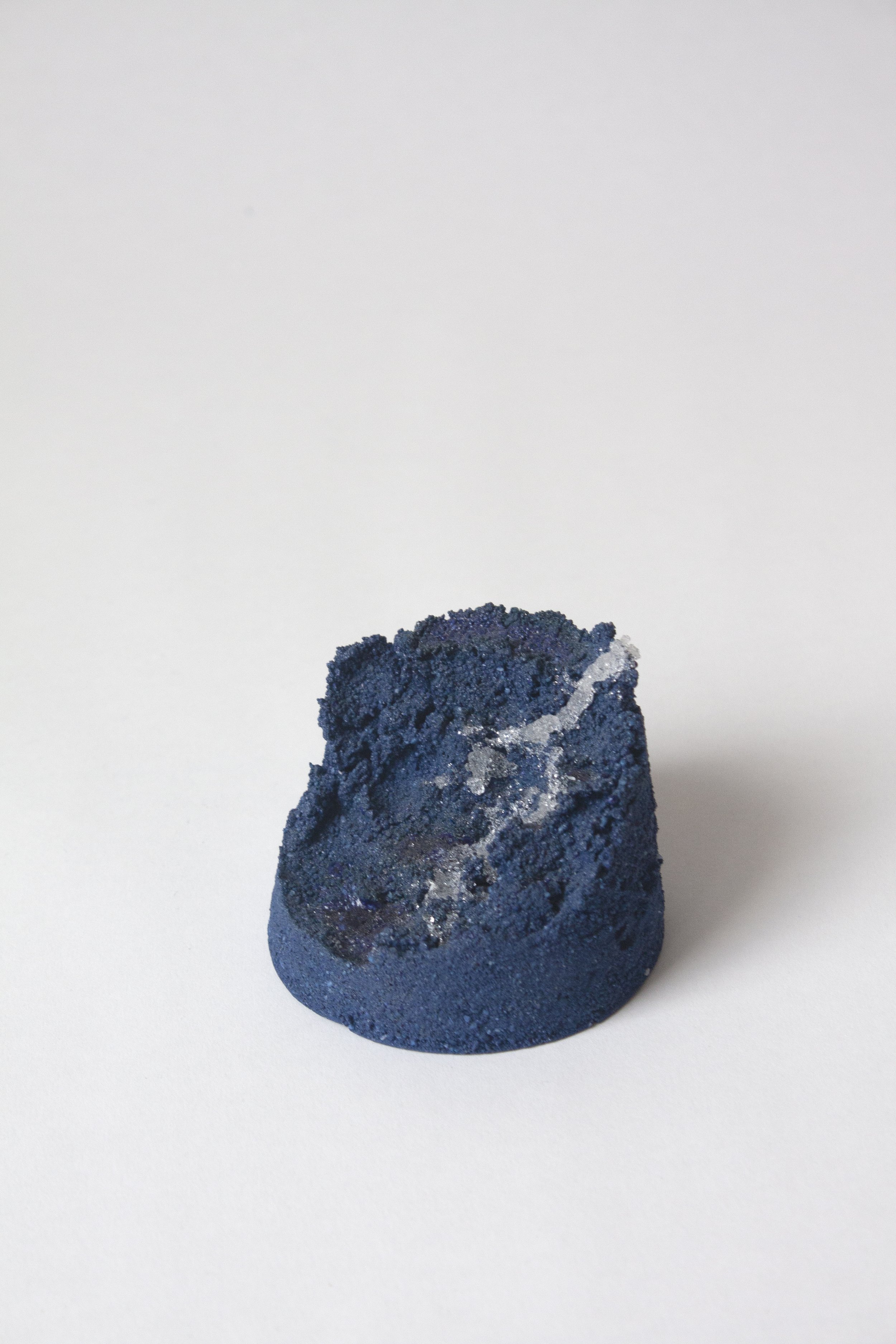 A small, dark-colored rock or mineral specimen with a rough, textured surface and shiny, metallic-looking patches, placed on a plain white background.