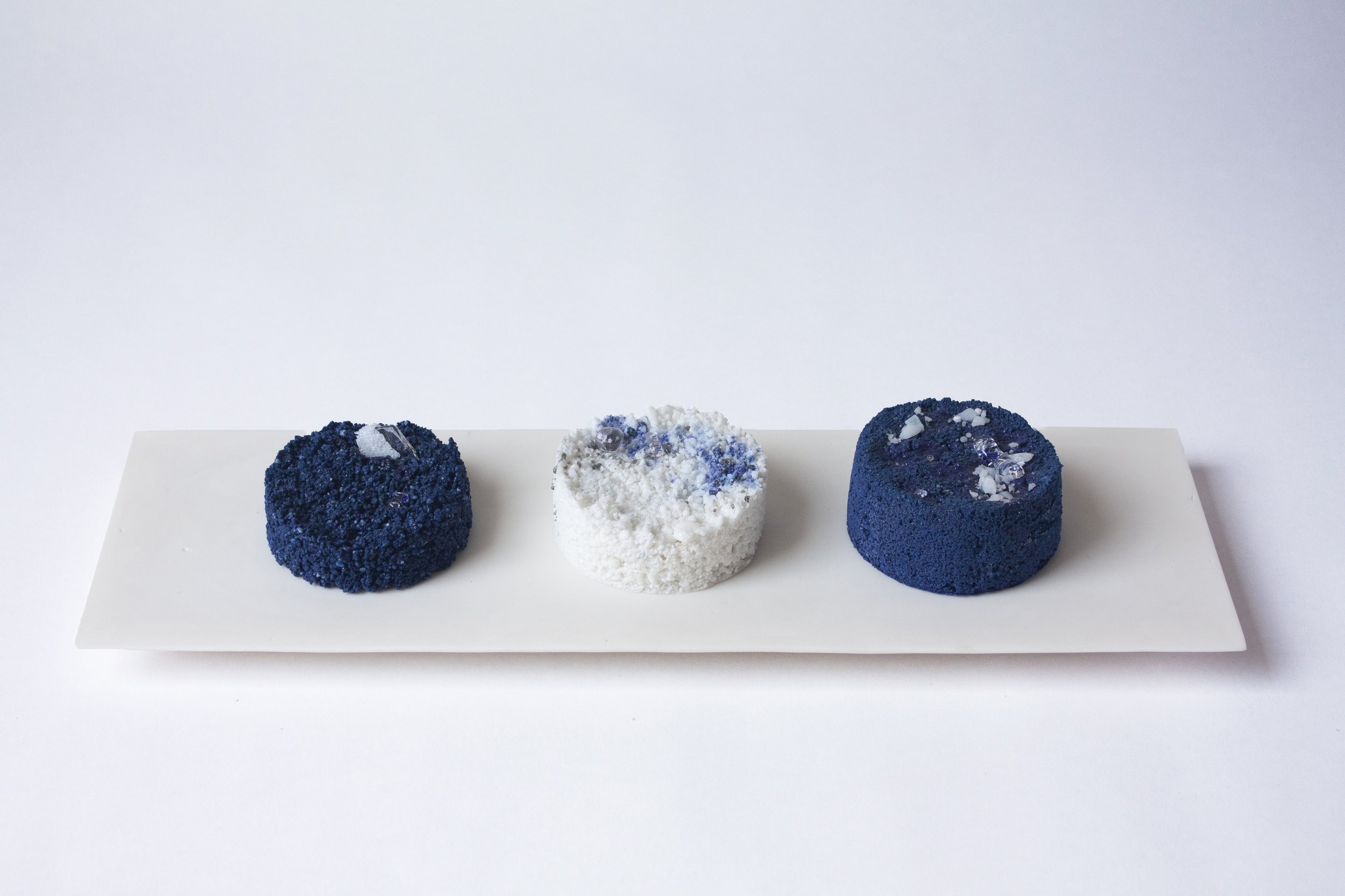 Three textured objects resembling donuts or cakes on a white rectangular plate against a plain white background, with the middle item being white and others dark blue with embellishments.