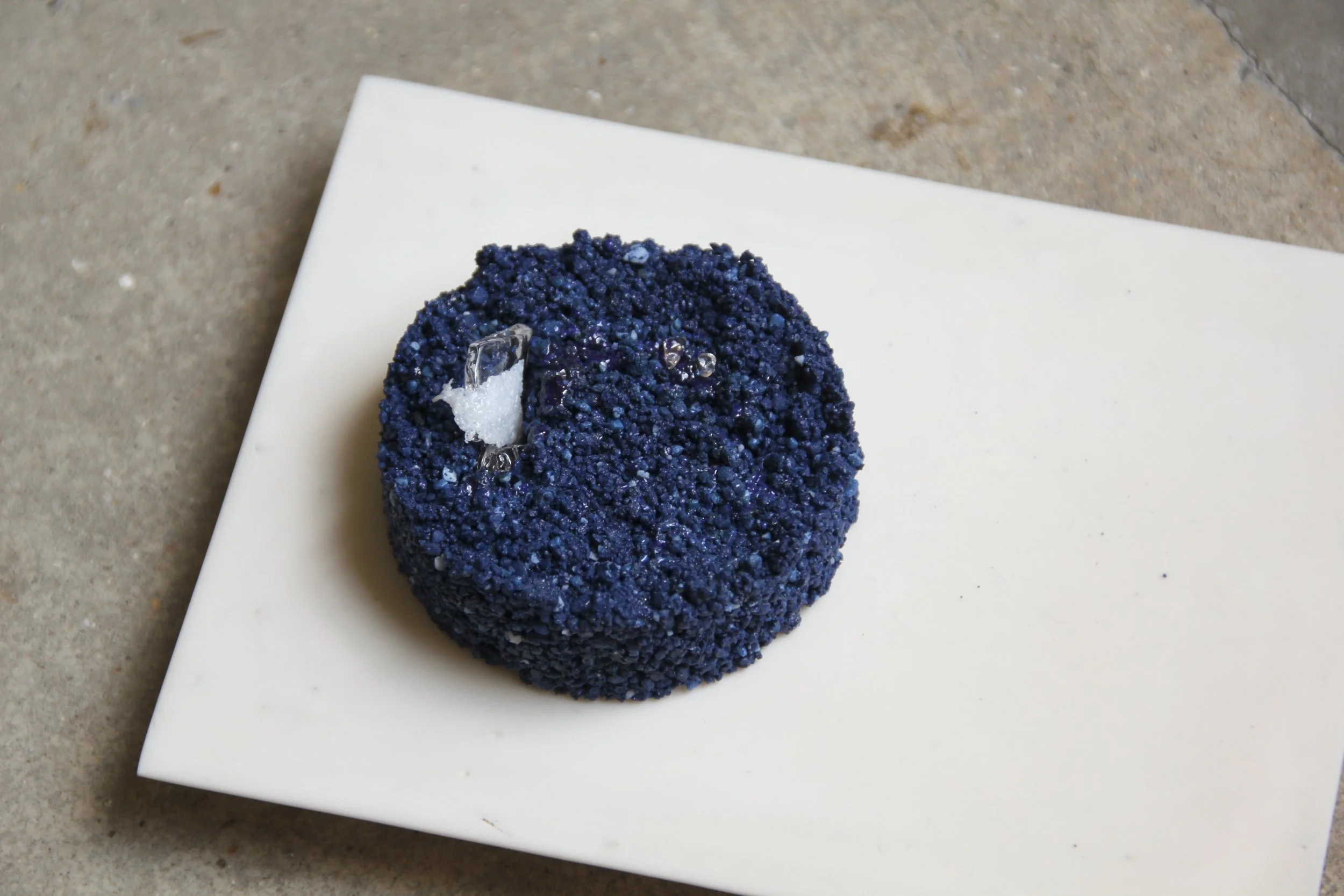 A blue, textured ring-looking sculpture with embedded clear glass pieces, placed on a white surface.