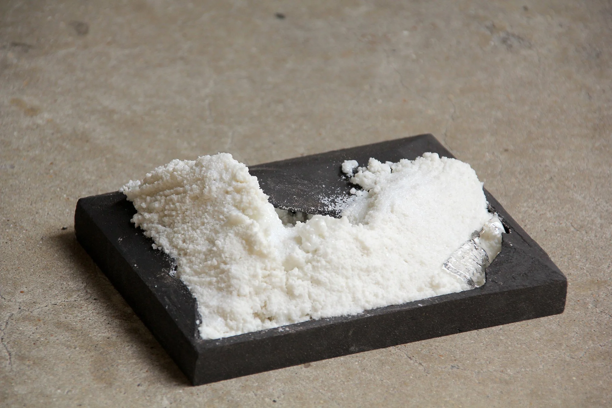 A black slate stone with white salt or powder on it, a metal spatula, and a roll of transparent plastic wrap on a textured gray surface.