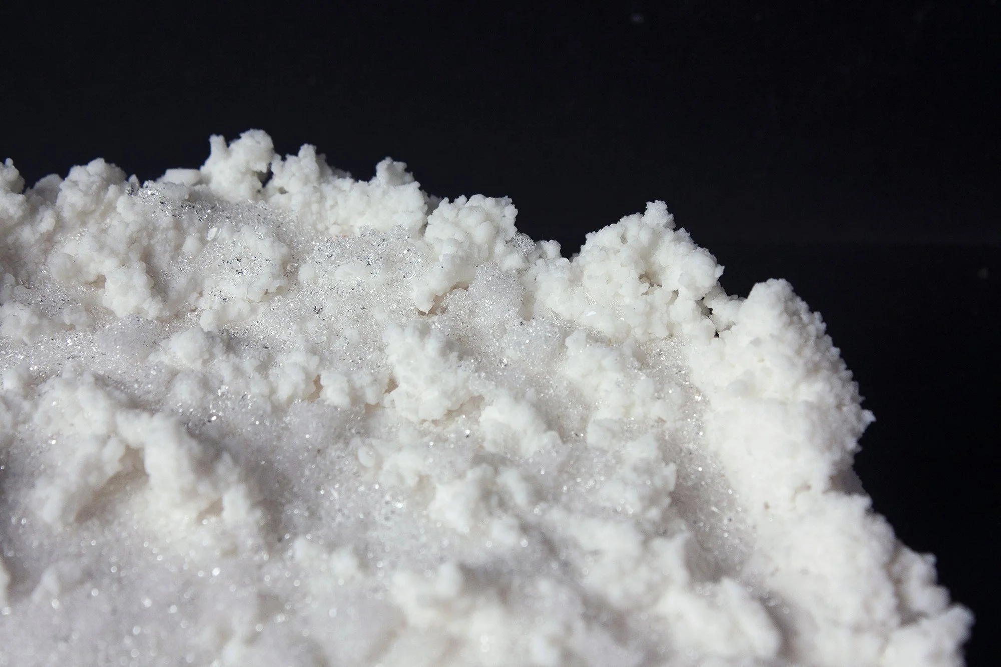 Close-up of white salt crystals with small silver-colored particles, set against a black background.
