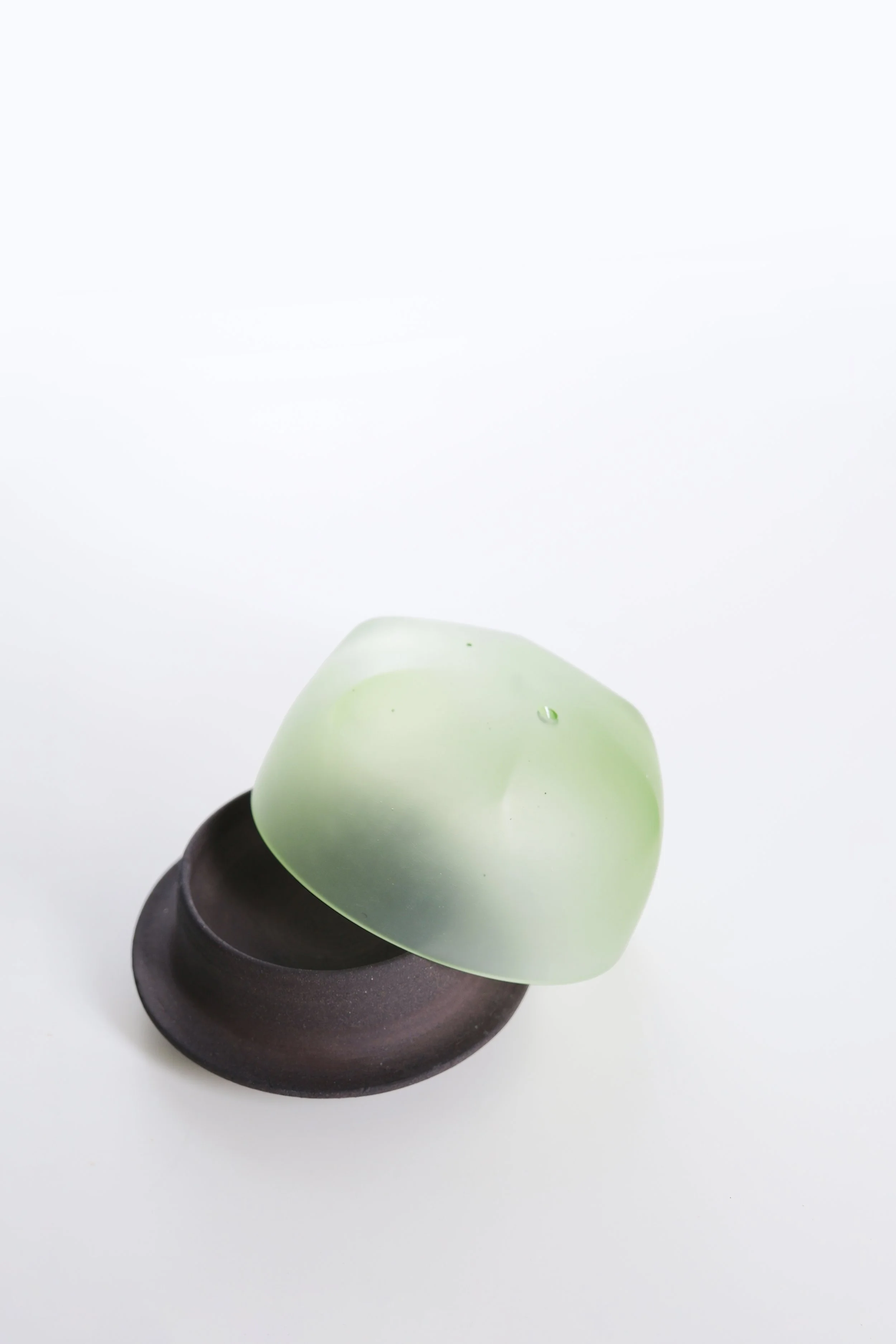 A green heart-shaped object with a water droplet on its surface, resting on a black circular base, against a plain white background.