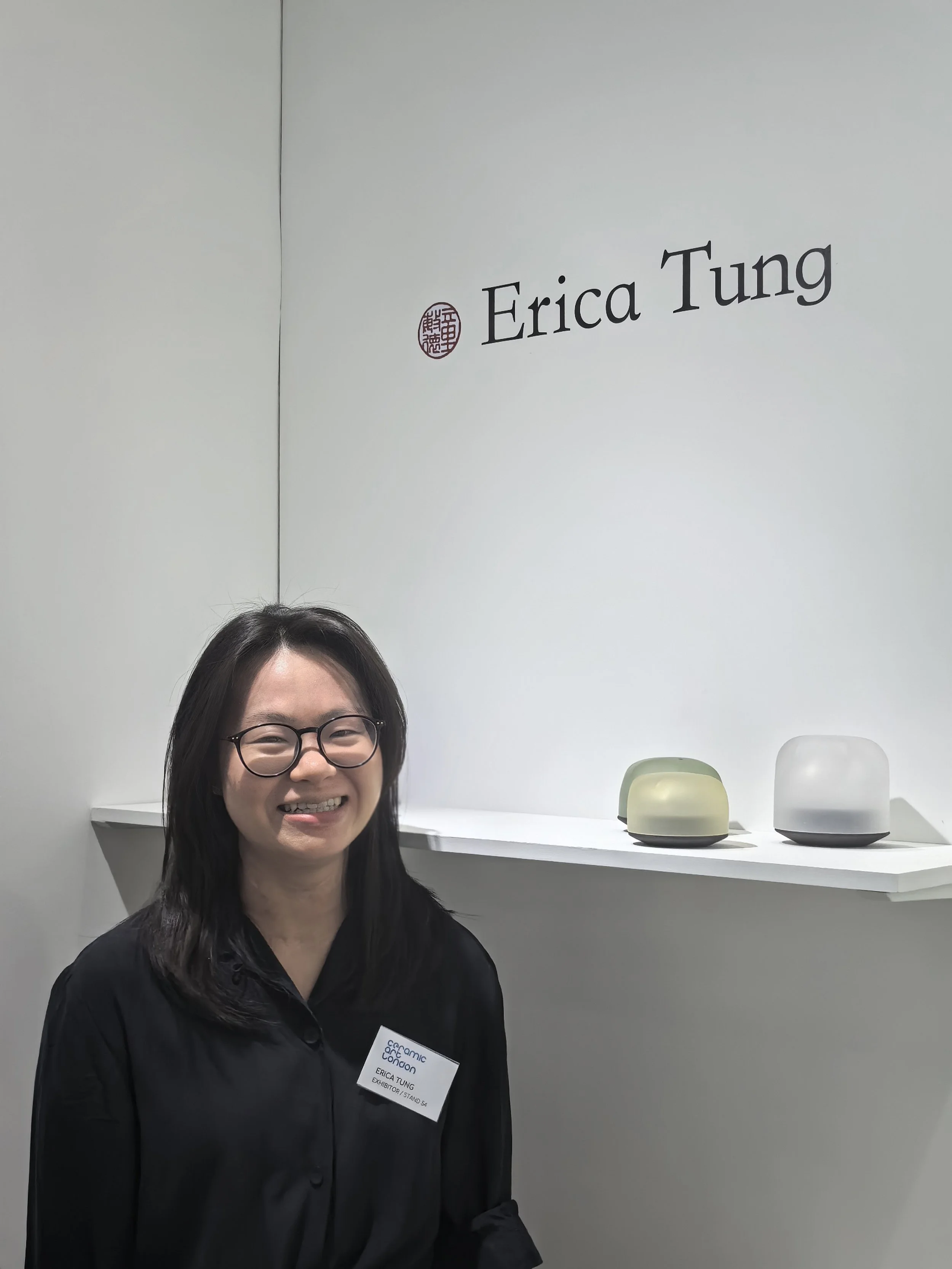 A woman with black hair and glasses smiling in front of a white wall with the name Erica Tung and three ceramic sculptures on a shelf.