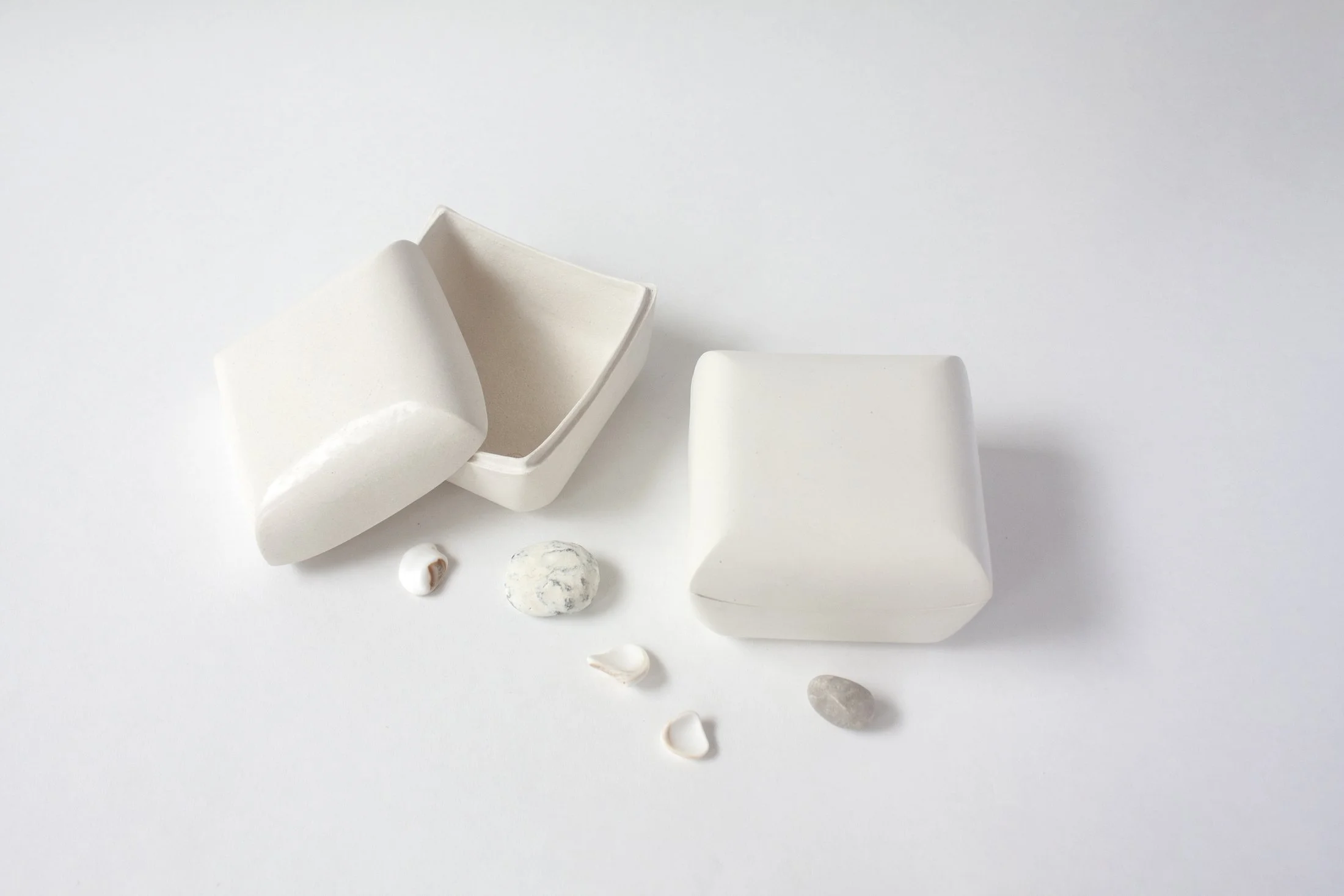 Various white ceramic objects, including a bowl, a rectangular block, and a curved piece, on a white background with scattered small stones and shells.