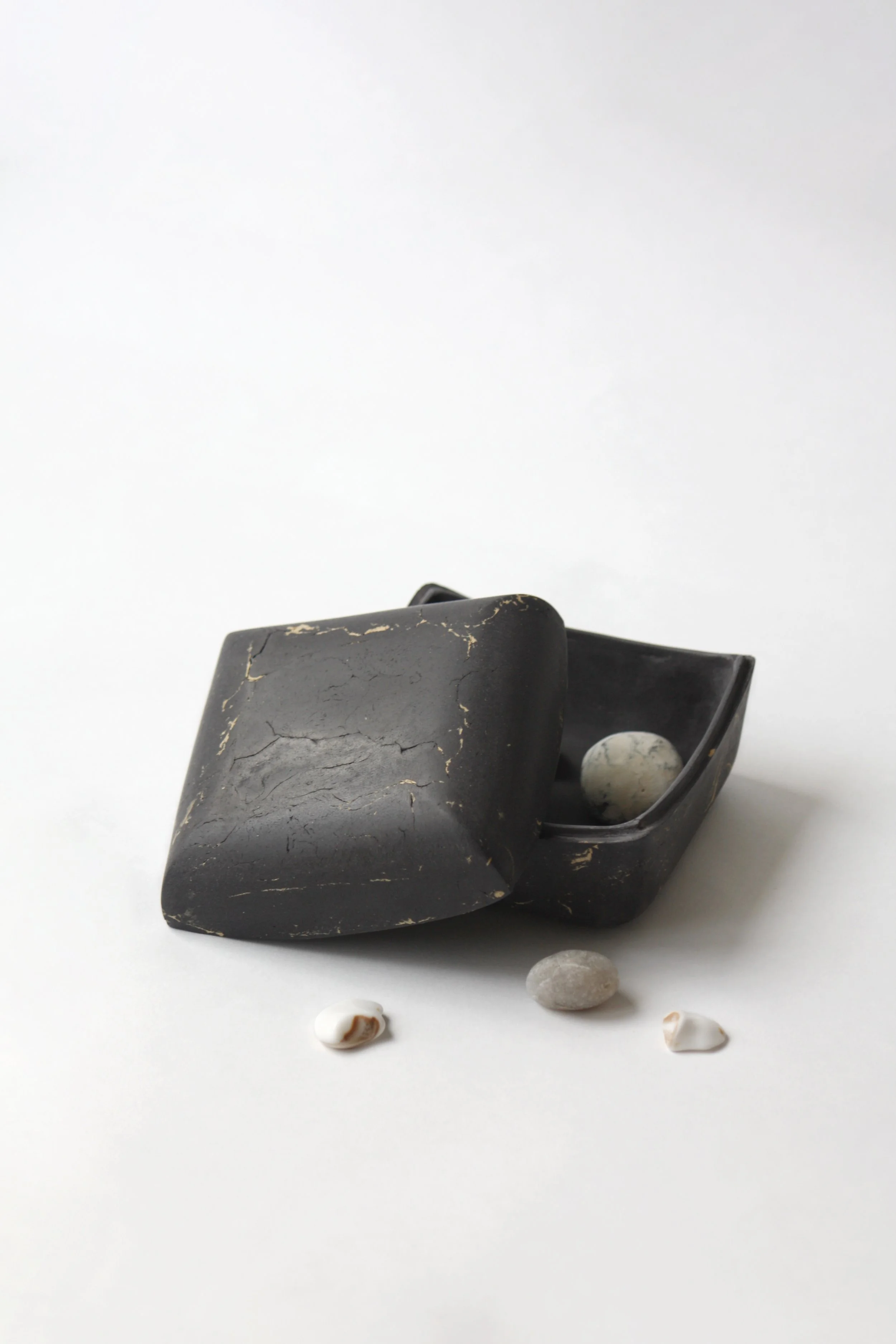 A black doorstop with a crackled surface, holding back a white object inside a black container, with small shells and stones scattered in front on a white surface against a plain white background.