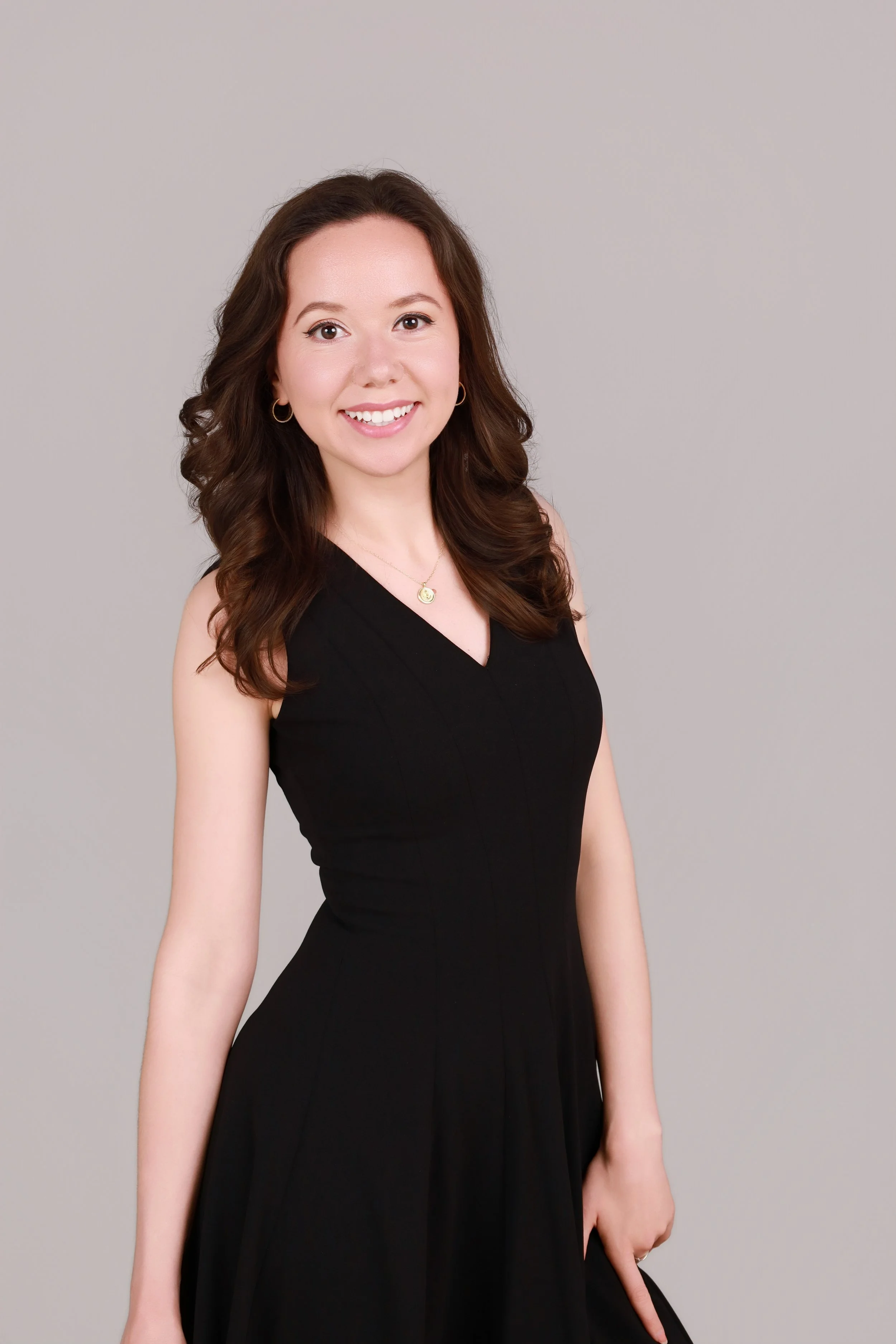 A woman with long, wavy brown hair, wearing a black sleeveless dress, gold hoop earrings, and a gold pendant necklace, smiling against a plain light gray background.