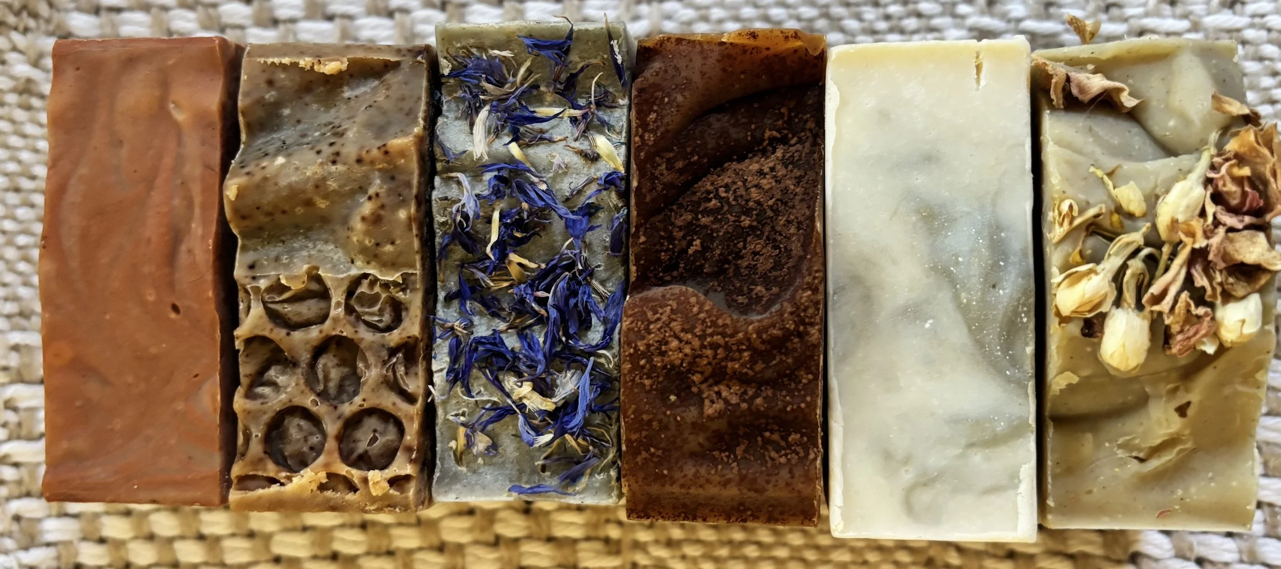 Six assorted handmade soap bars arranged in a row on a woven surface, each with different textures and colors handmade by Skipwillow Farm