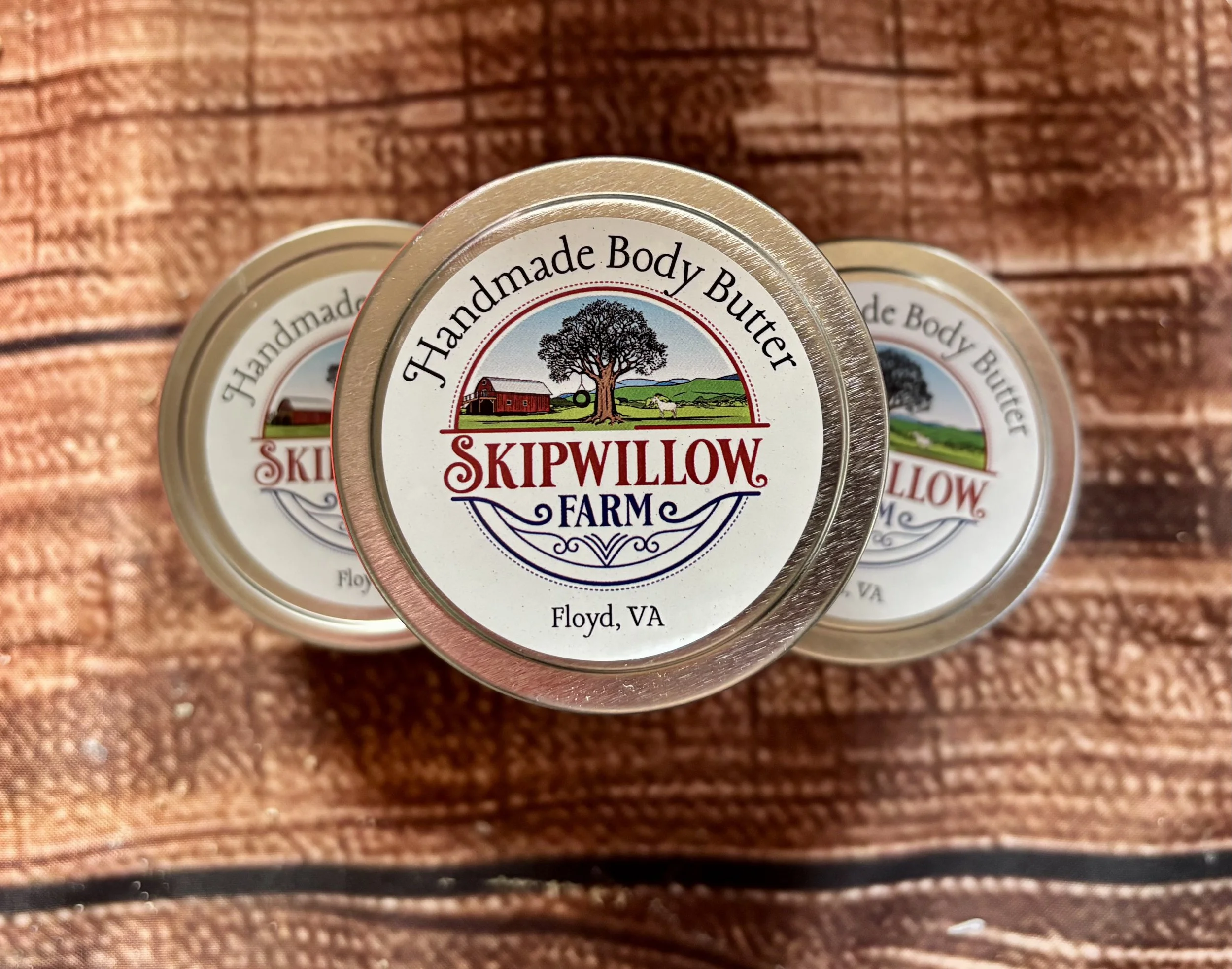 Close-up of a cosmetic container labeled 'Handmade Body Butter' from Skipwillow Farm in Floyd, VA, with a rustic wooden surface background.