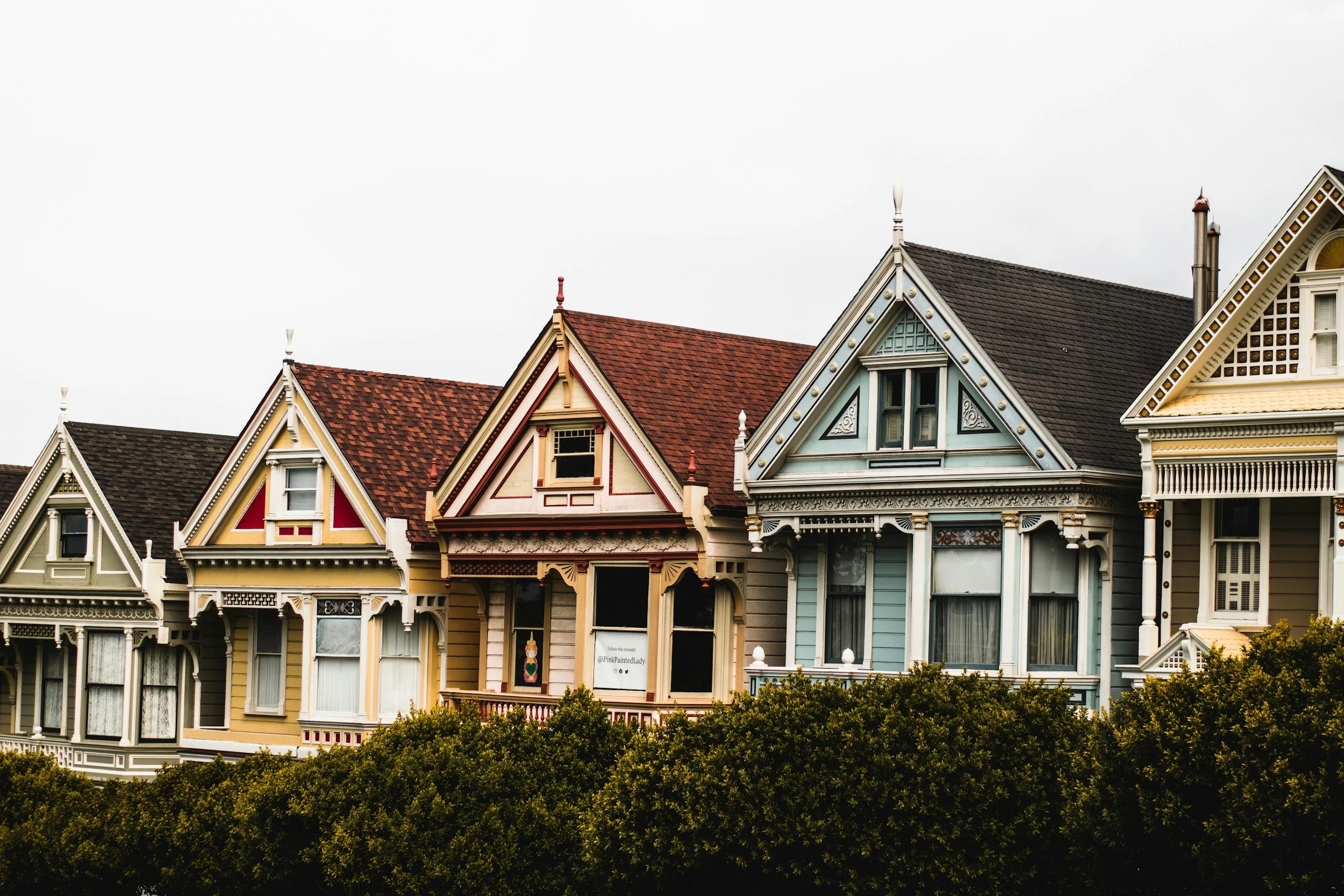 Colorful Victorian-style houses with detailed woodwork and gabled roofs in a row, partially obscured by green shrubbery in the foreground.