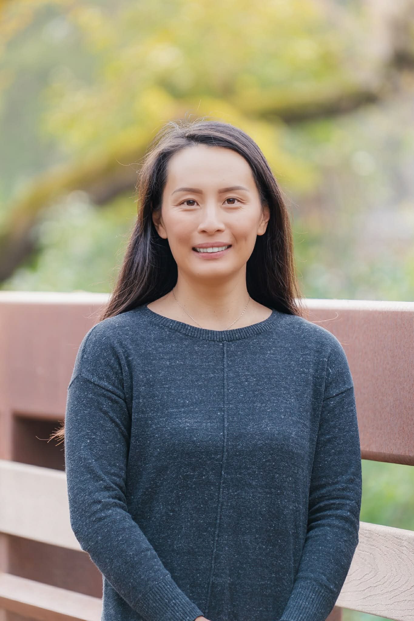 A woman with long dark hair wearing a dark blue sweater standing outdoors by a wooden railing with trees and greenery in the background.