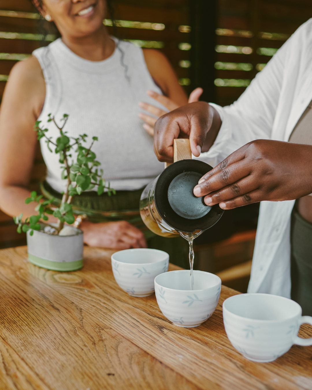 Person pouring tea into cups on a wooden table while another person looks on smiling, with a potted plant nearby.
