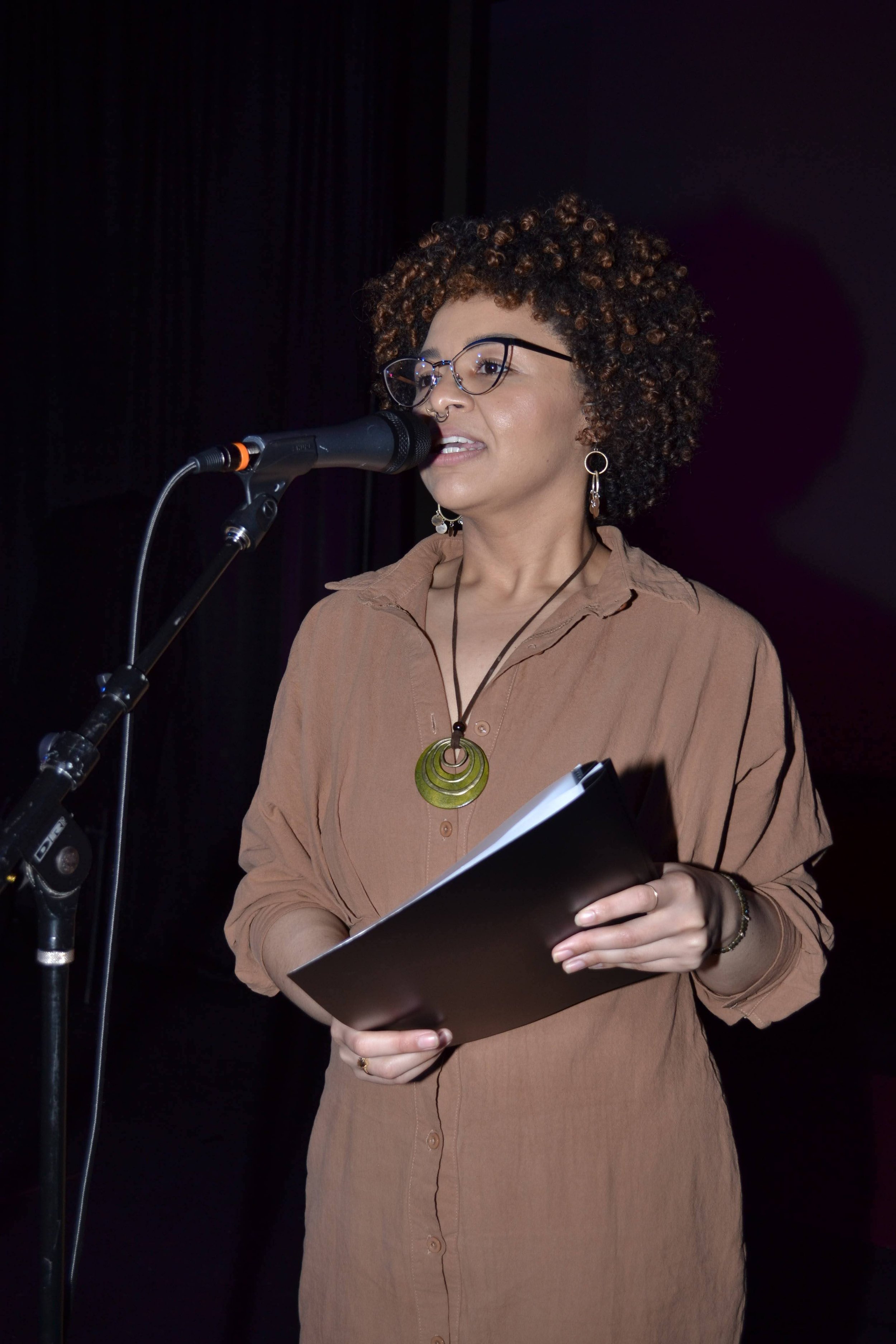 Mars Mena standing in front of a microphone, holding a closed notebook or folder, and speaking.