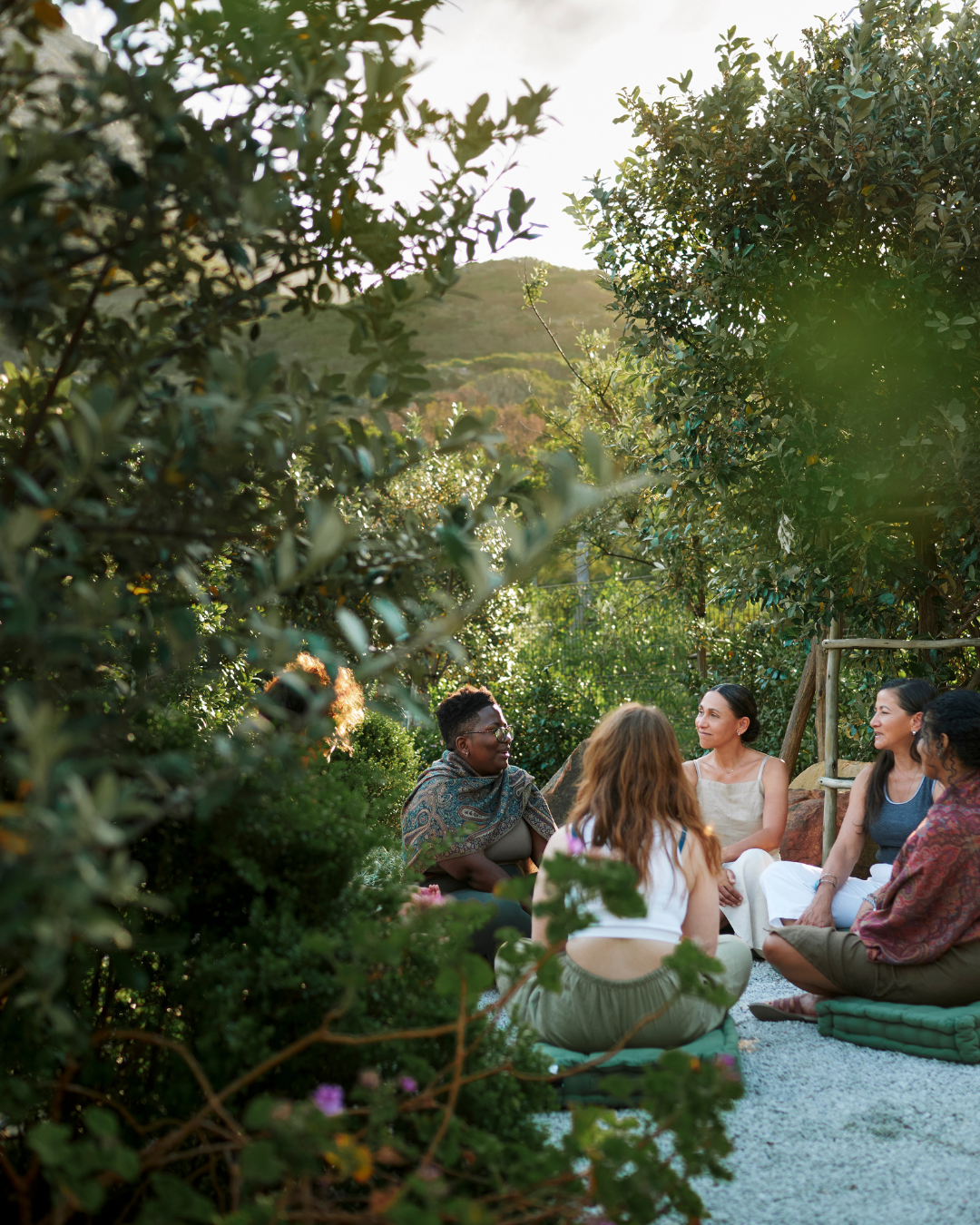 A group of women sitting on cushions outdoors, engaged in conversation among lush greenery and trees, with a hill in the background.