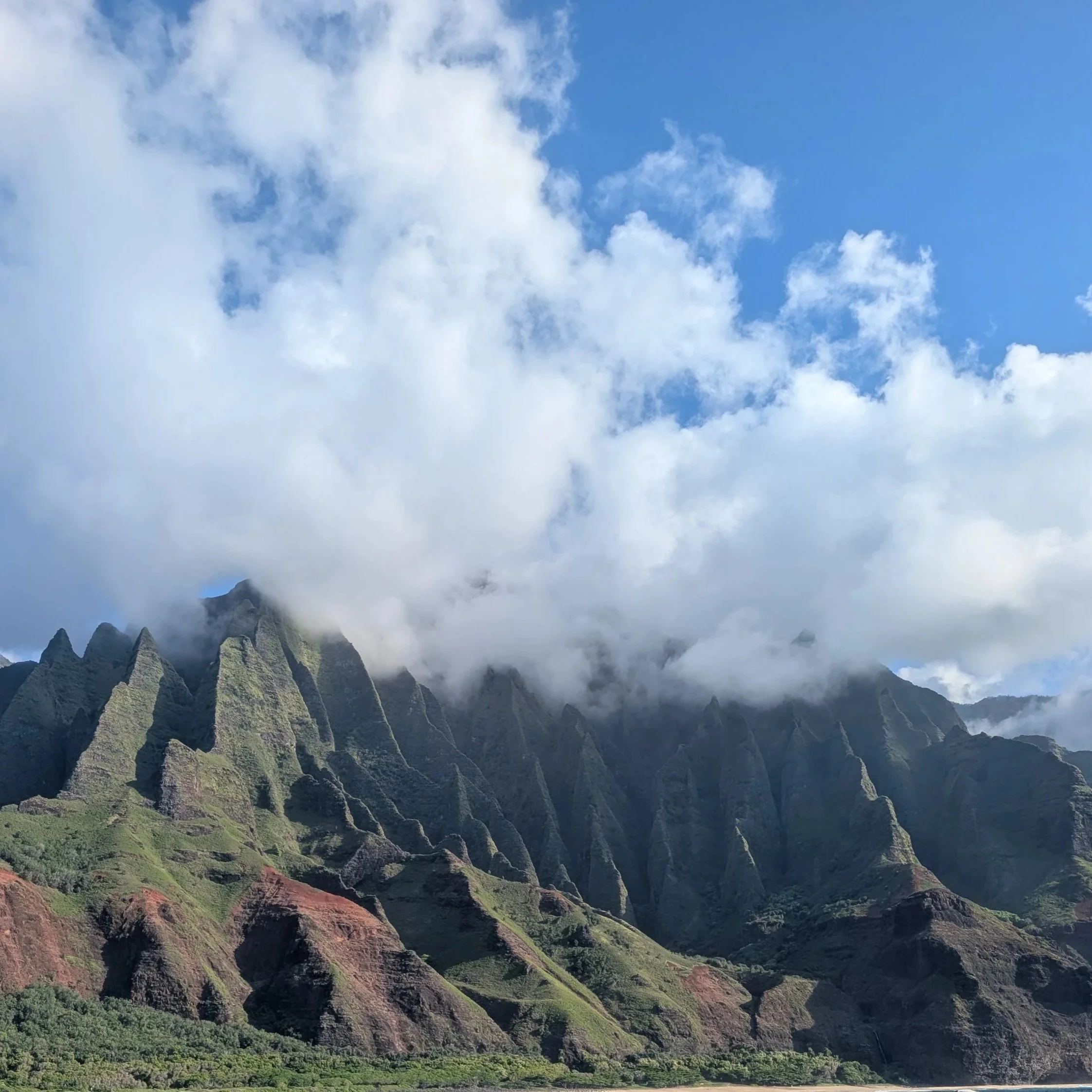 Towering green and brown mountains partly covered by clouds with a lush green valley below. Taken Nā Pali Coast, Kauai, Hawaii.