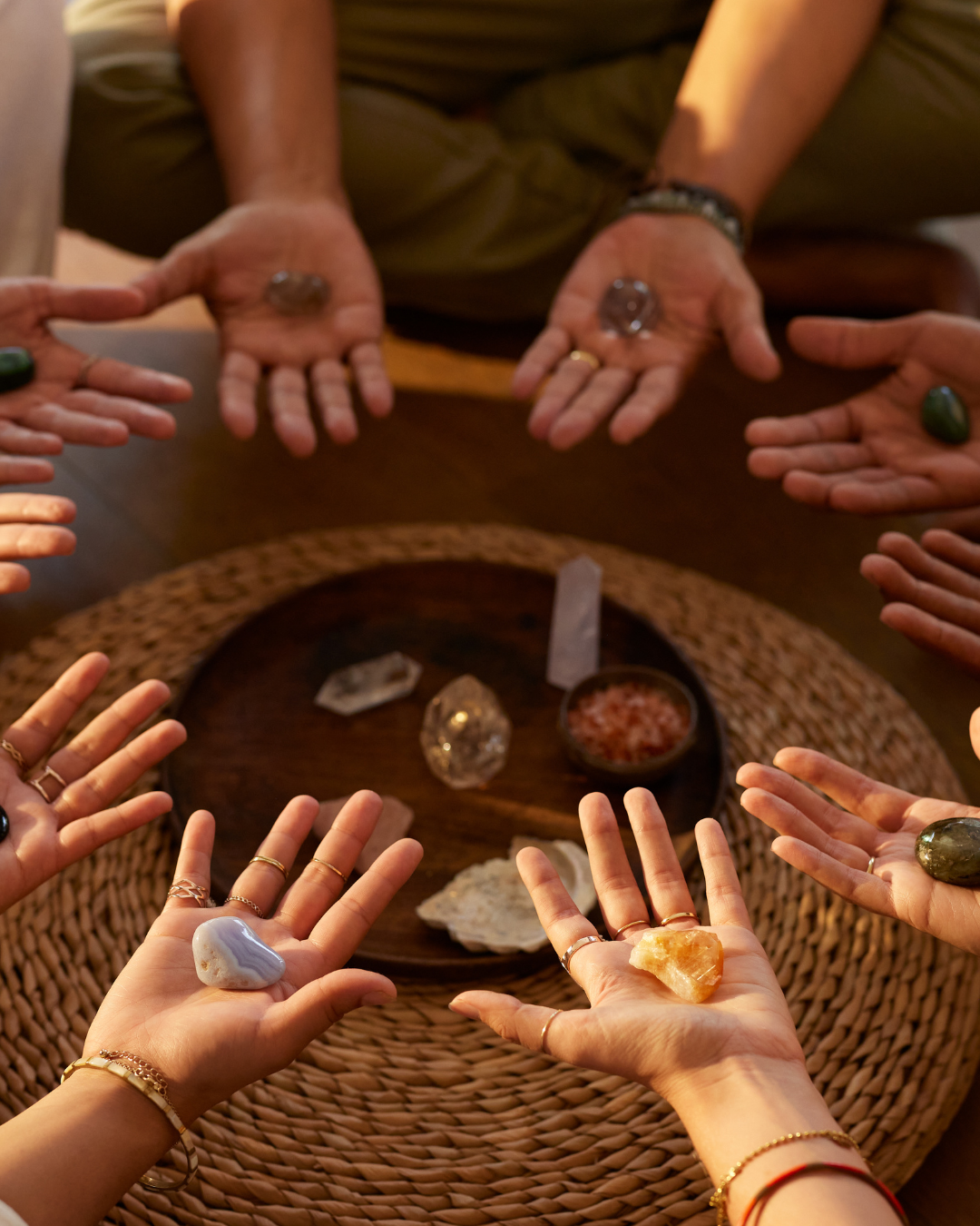 A group of people sitting around a table holding various crystals and stones in their hands, with more crystals, stones, and salt on a round wooden tray in the center.
