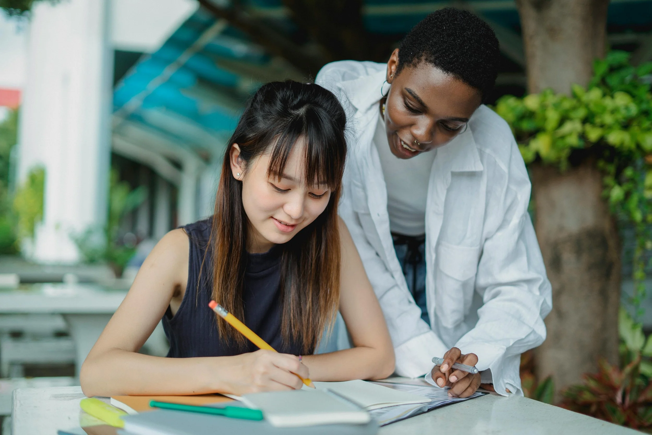 A young woman with long dark hair and a young man with short curly hair are outside, leaning over a table and working on a project together. The woman is holding a pencil, and the man is looking at her work, smiling. There are notebooks and colorful pens on the table.