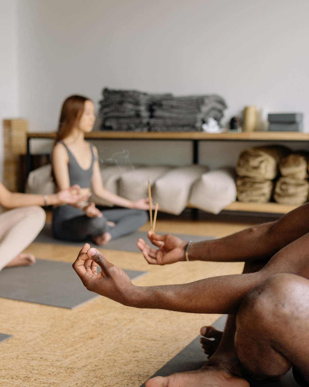 People practicing meditation in a room, with a person holding two incense sticks and the others sitting on yoga mats in the background.