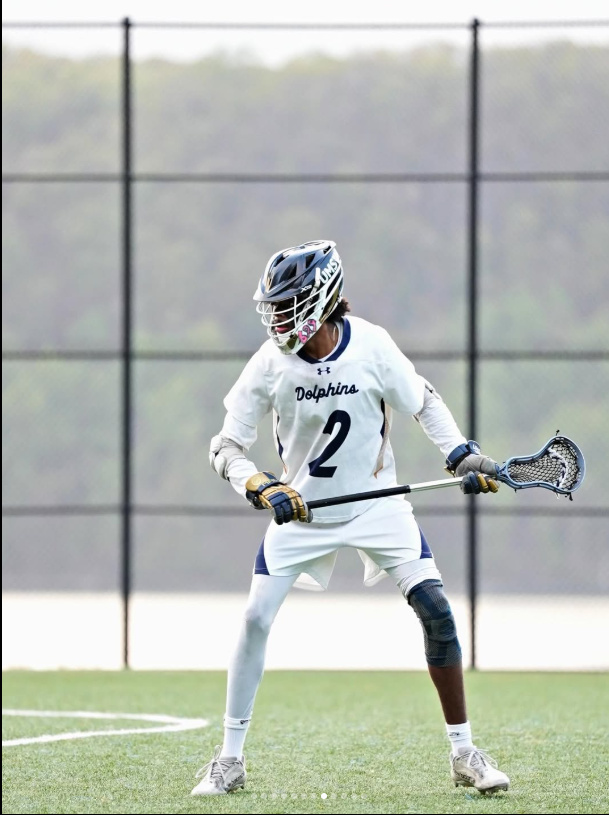 Lacrosse player in white uniform with the number 2 and 'Dolphins' on the jersey, holding a lacrosse stick on a field.