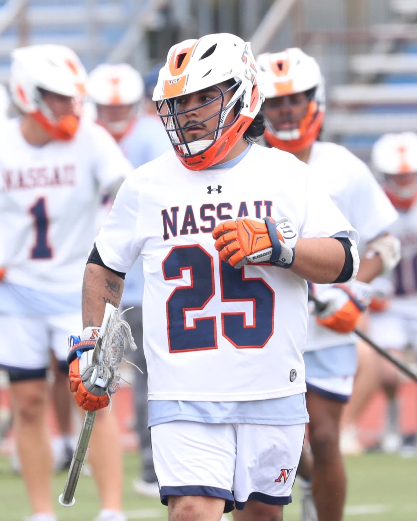 Lacrosse player in white jersey with 'NASSAU' written on it, number 25, wearing helmet and gloves, holding lacrosse stick, standing on the field with teammates in the background.