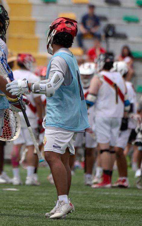 A young baseball or lacrosse player wearing a red helmet with face protection, a light blue vest, white shorts, and athletic shoes, standing on a sports field during a game, with other players and spectators in the background.