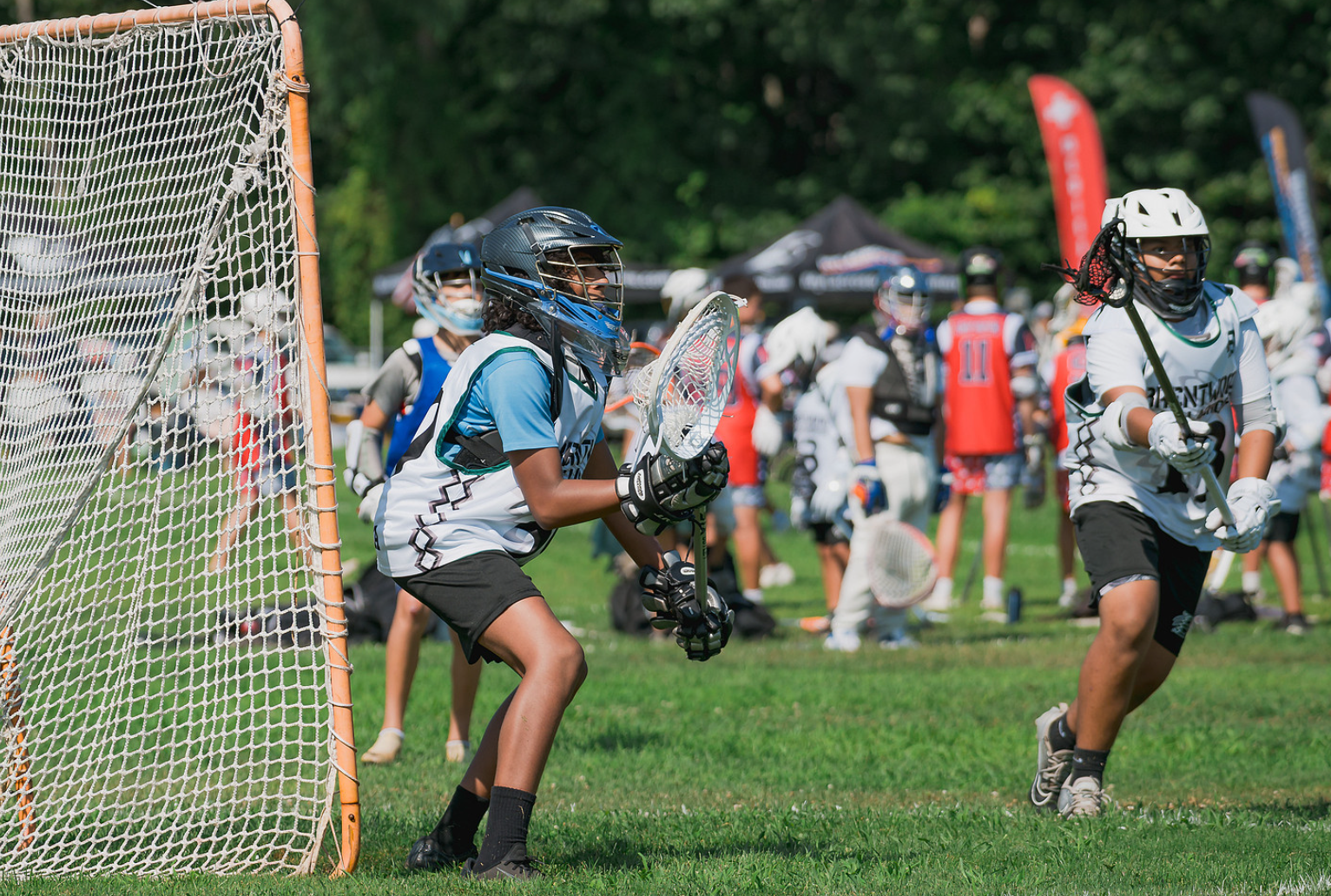 Lacrosse players wearing helmets and gear practicing on a grassy field during a game with spectators and tents in the background.