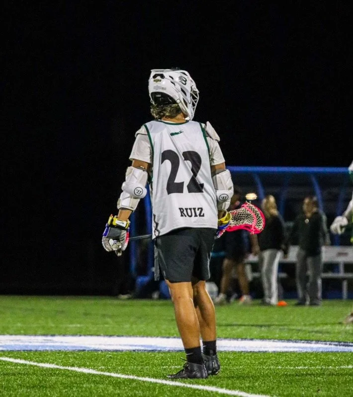 Lacrosse player wearing a helmet, gloves, and a jersey with the number 22 and the name RUIZ, holding a lacrosse stick, standing on a field at night.