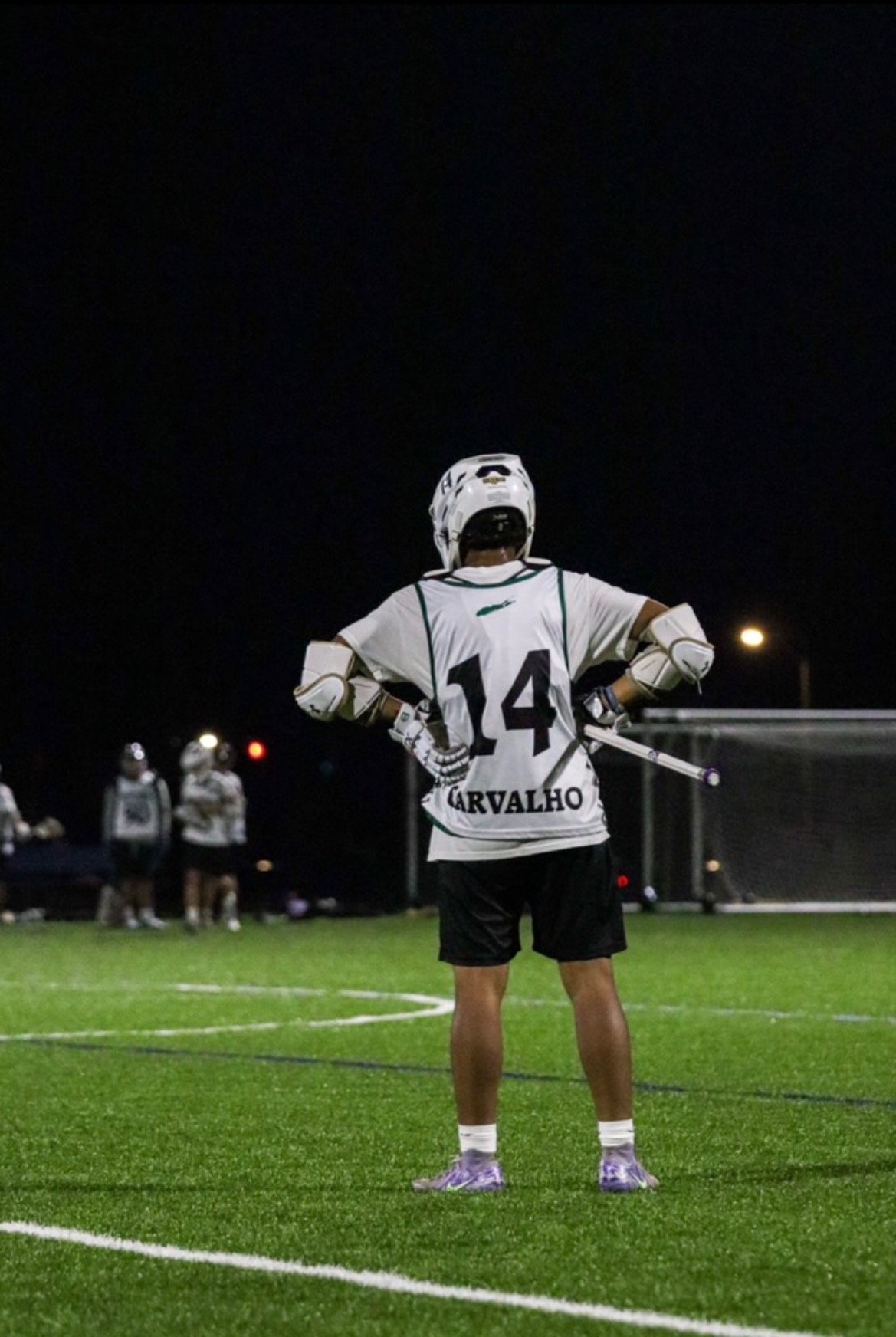 A hockey player standing on a field at night, wearing a white jersey with the number 14 and the name 'Carvalho' on the back, holding a stick, with other players in the background.
