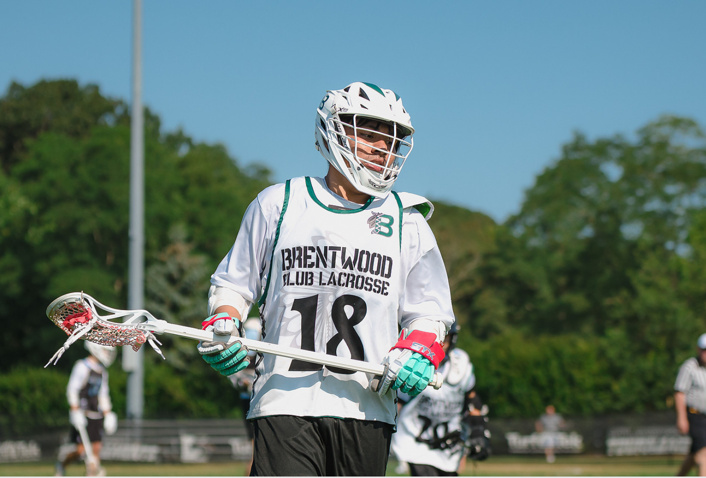 Lacrosse player wearing a helmet, white jersey with 'Brentwood Club Lacrosse' and number 18, holding a lacrosse stick, on a field with trees in the background.
