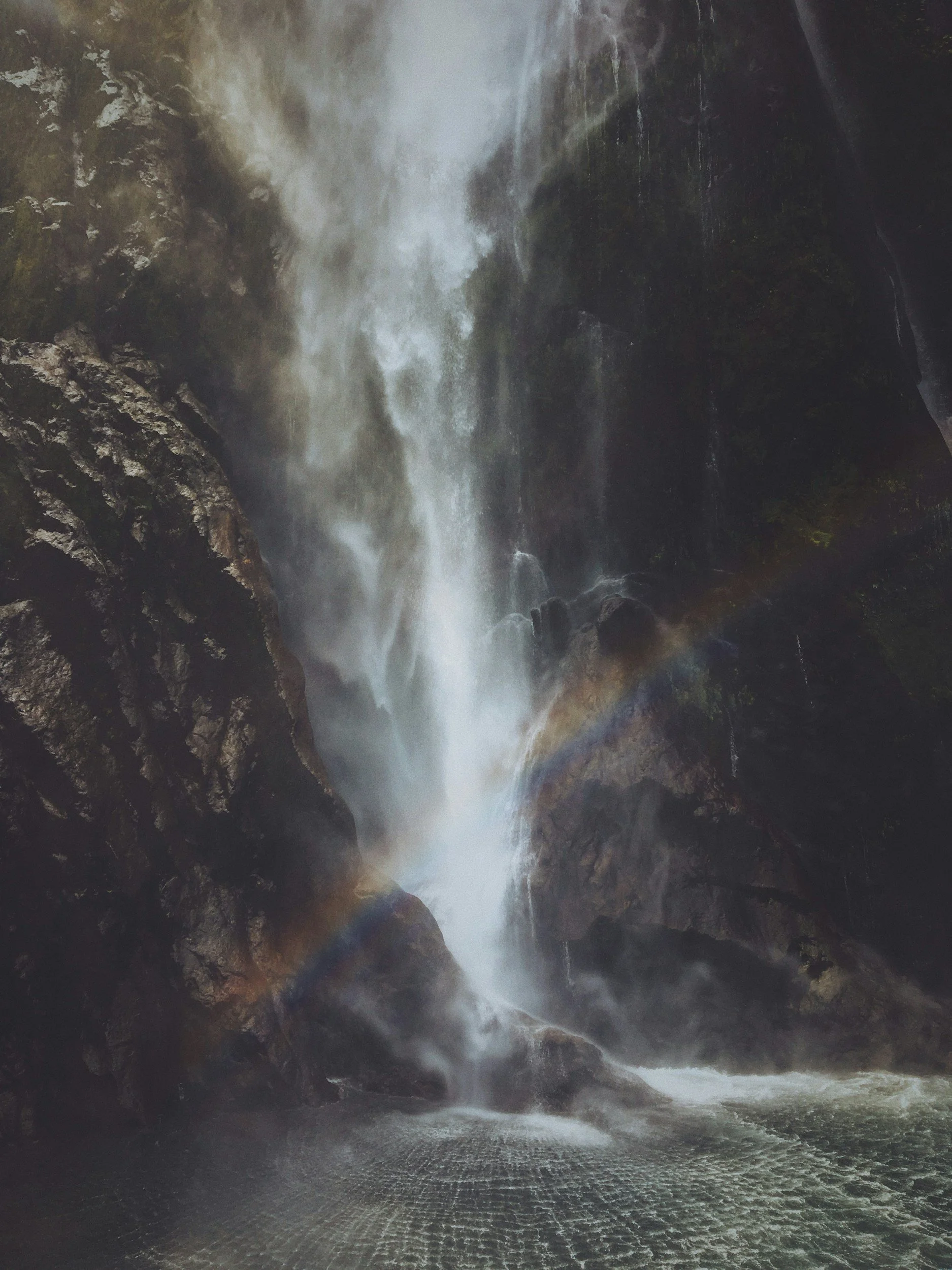 A powerful waterfall cascading down dark rocks, creating mist at the base and a rainbow arching over the lower part of the waterfall.