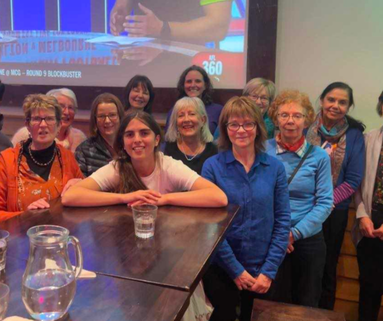 Group of women and young girl posing for a photo at a social gathering indoors, with a large screen in the background showing a game or broadcast.