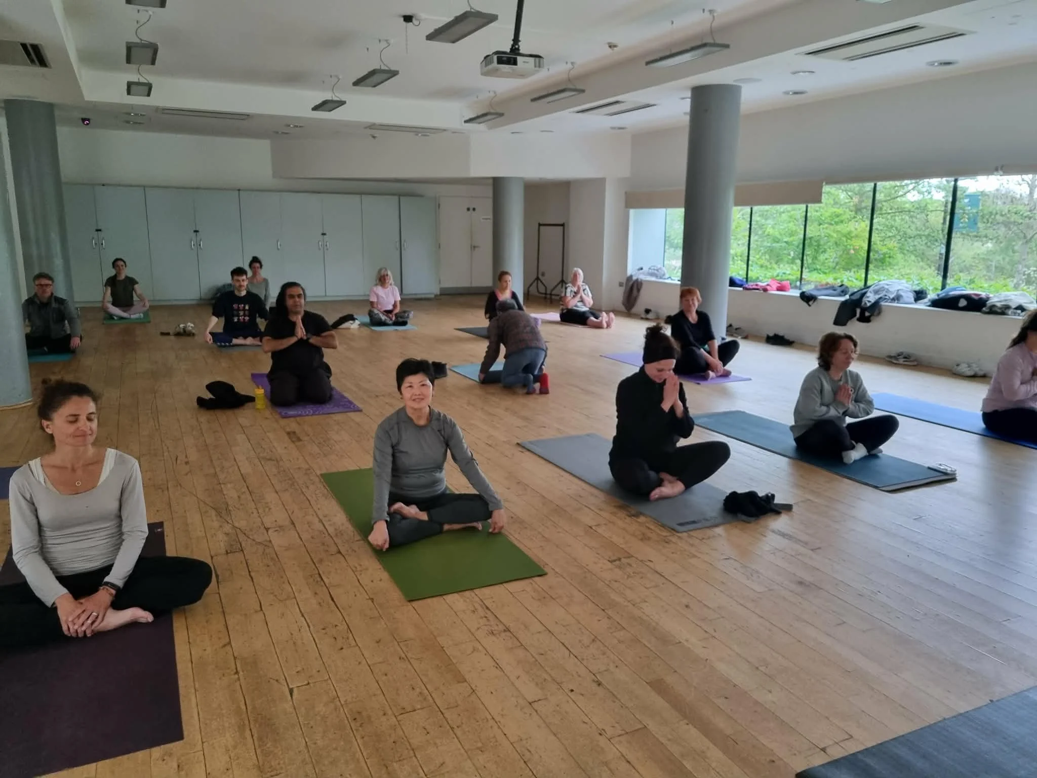 Group of people participating in a yoga or meditation class in a bright indoor studio, sitting cross-legged on mats, some with palms pressed together in a prayer position.