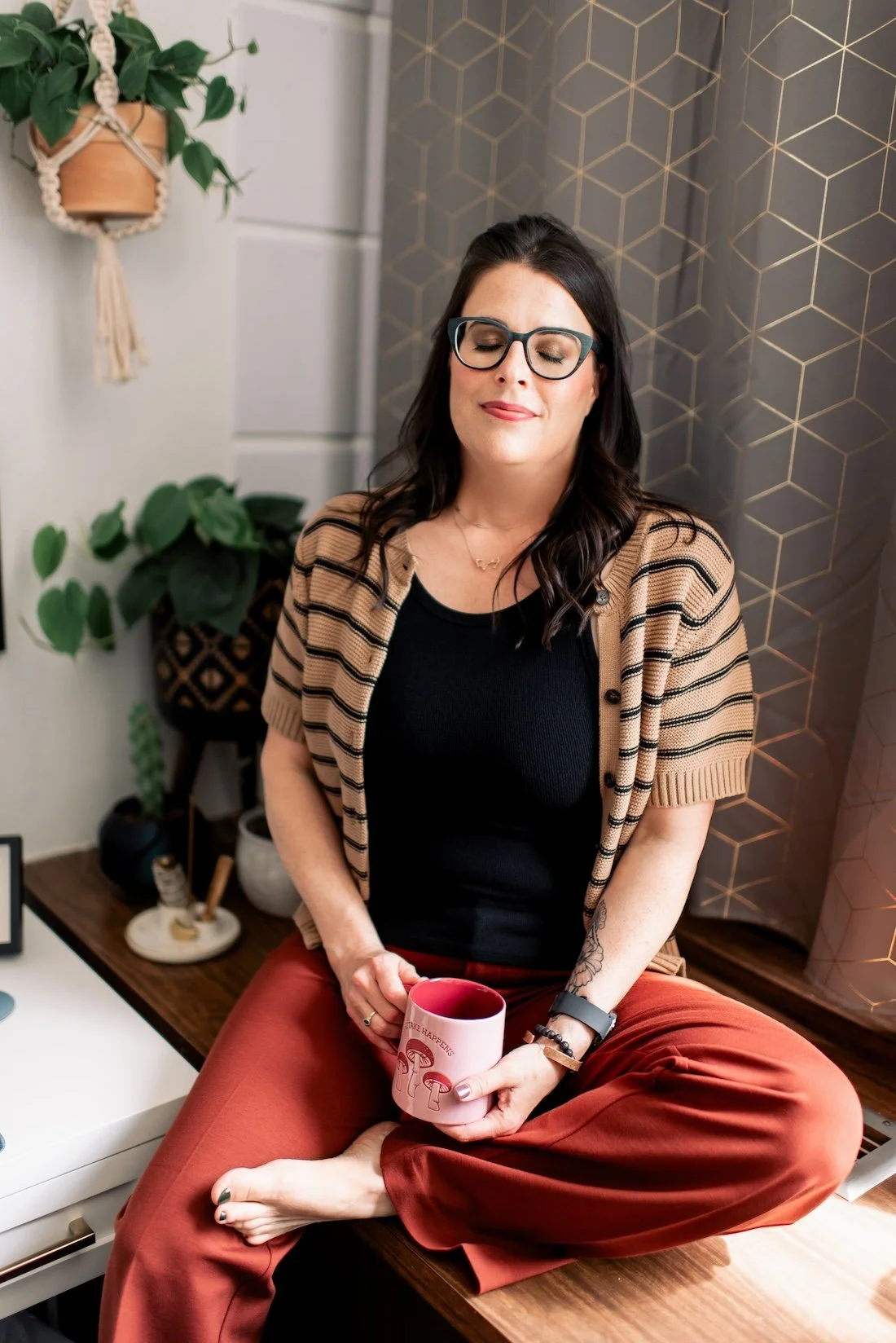 A woman with dark hair, glasses, and tattoos sitting cross-legged on a wooden surface, holding a pink mug with tea or coffee, in a cozy indoor space with plants and decorative elements.