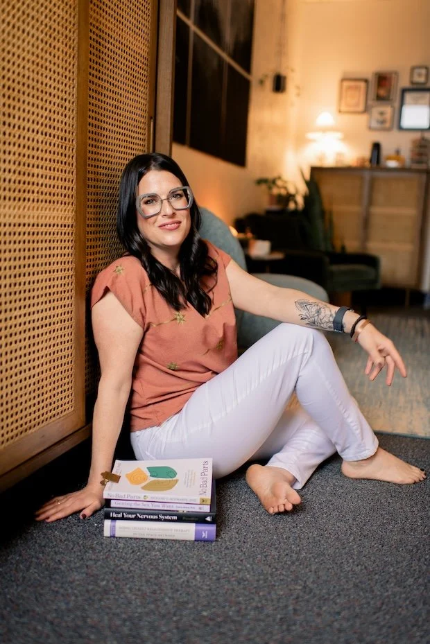 Joann Chevaillier, LPC-Associate, sitting comfortably on the floor in a cozy room during a Psychotherapy session, leaning against a wooden wall with a stack of books next to her.