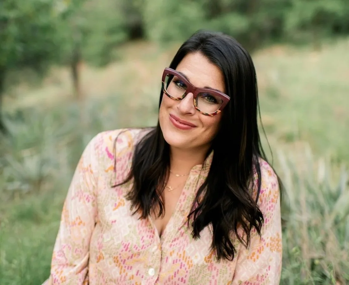 A woman with long dark hair and glasses smiling outdoors in a natural setting with greenery in the background.