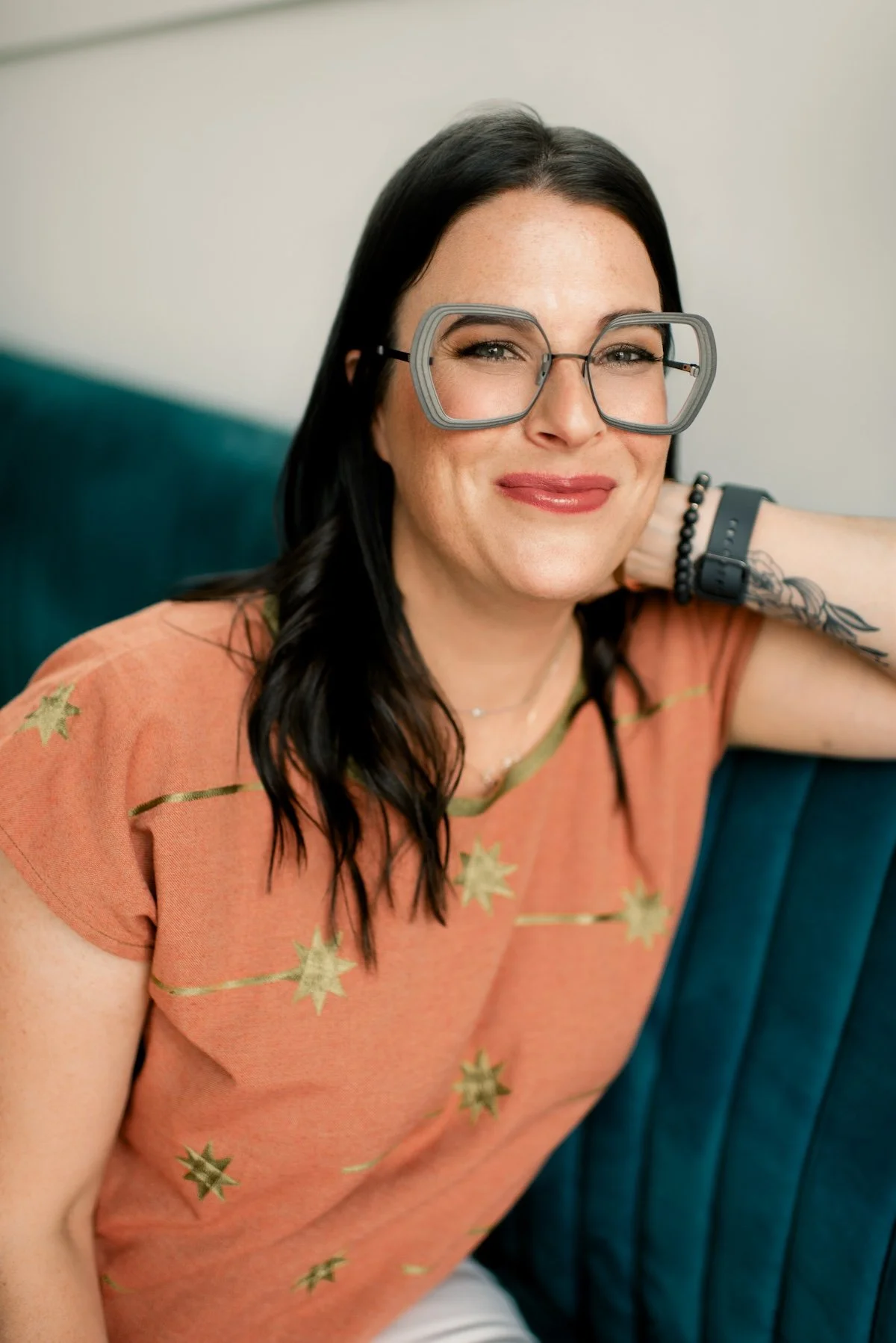 Meet your therapist, Joann Chevaillier, LPC-Associate. Joann sits smiling on a dark teal couch wearing glasses and a peach colored shirt.